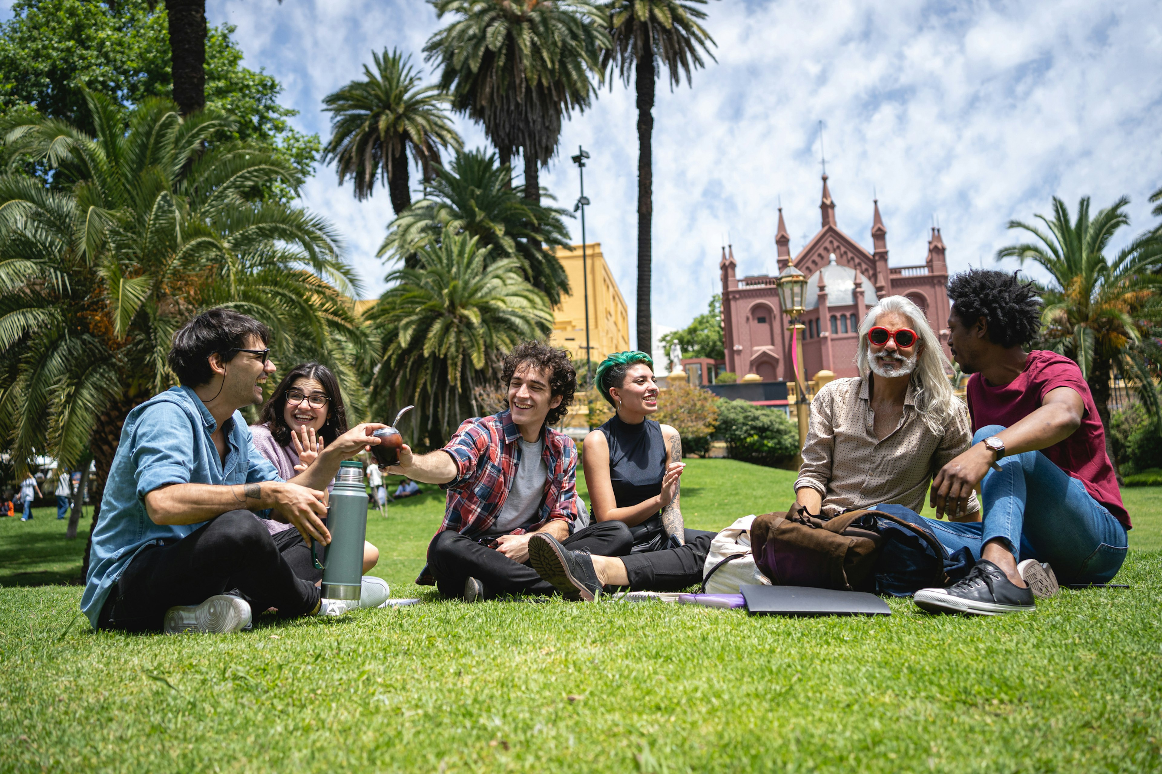 A group of students sit on the grass and chat in a park in the sunshine. One holds a thermos and is passing a gourd of mate to another