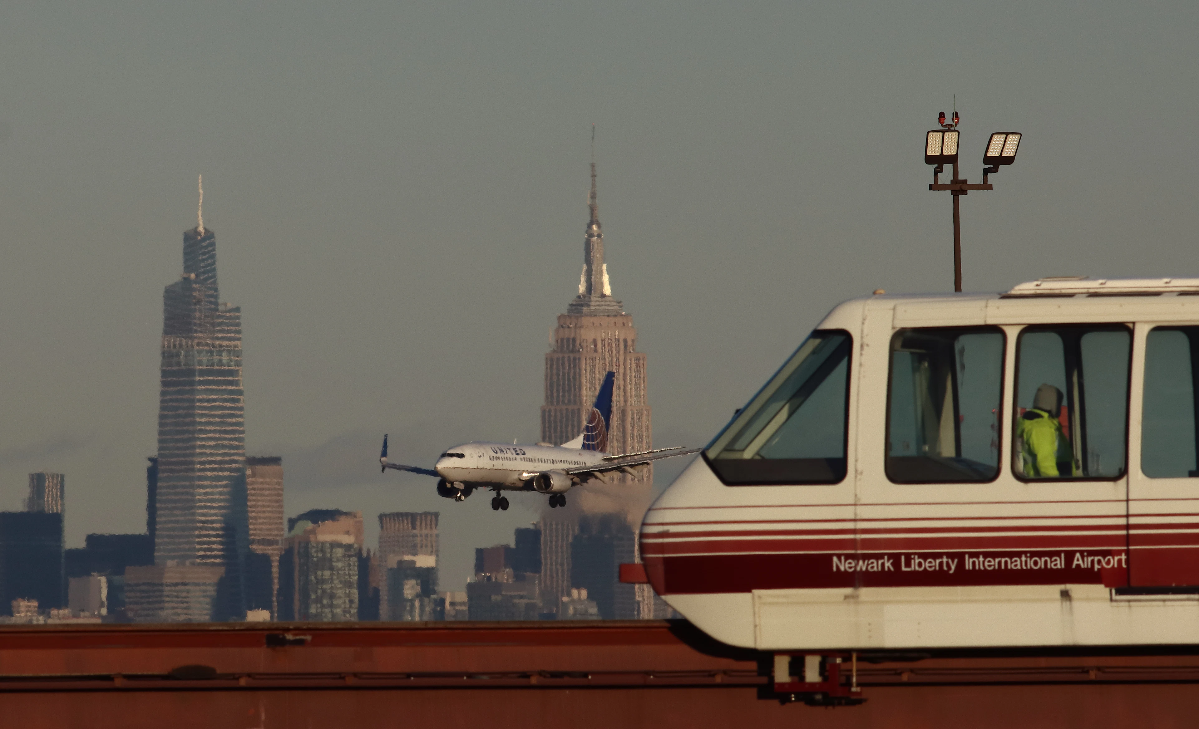 Самолет United Airlines летит перед зданием Эмпайр Стейт Билдинг и One Vanderbilt в Нью-Йорке, когда он приземляется, пока AirTrain проходит мимо, аэропорт Ньюарк Либерти, Ньюарк, Нью-Джерси, США