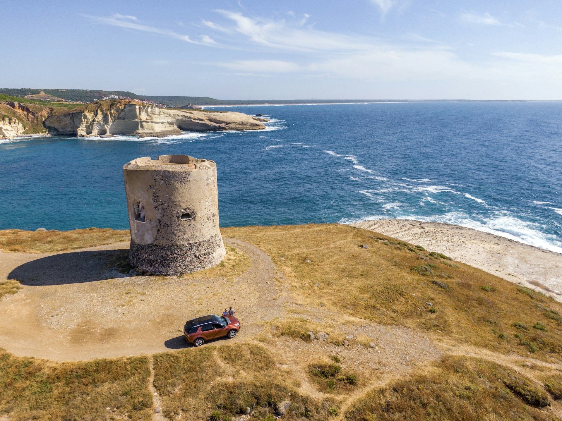 A red car sits in front of a beautiful bay with a ruined tower in summertime, Sardinia, Italy.