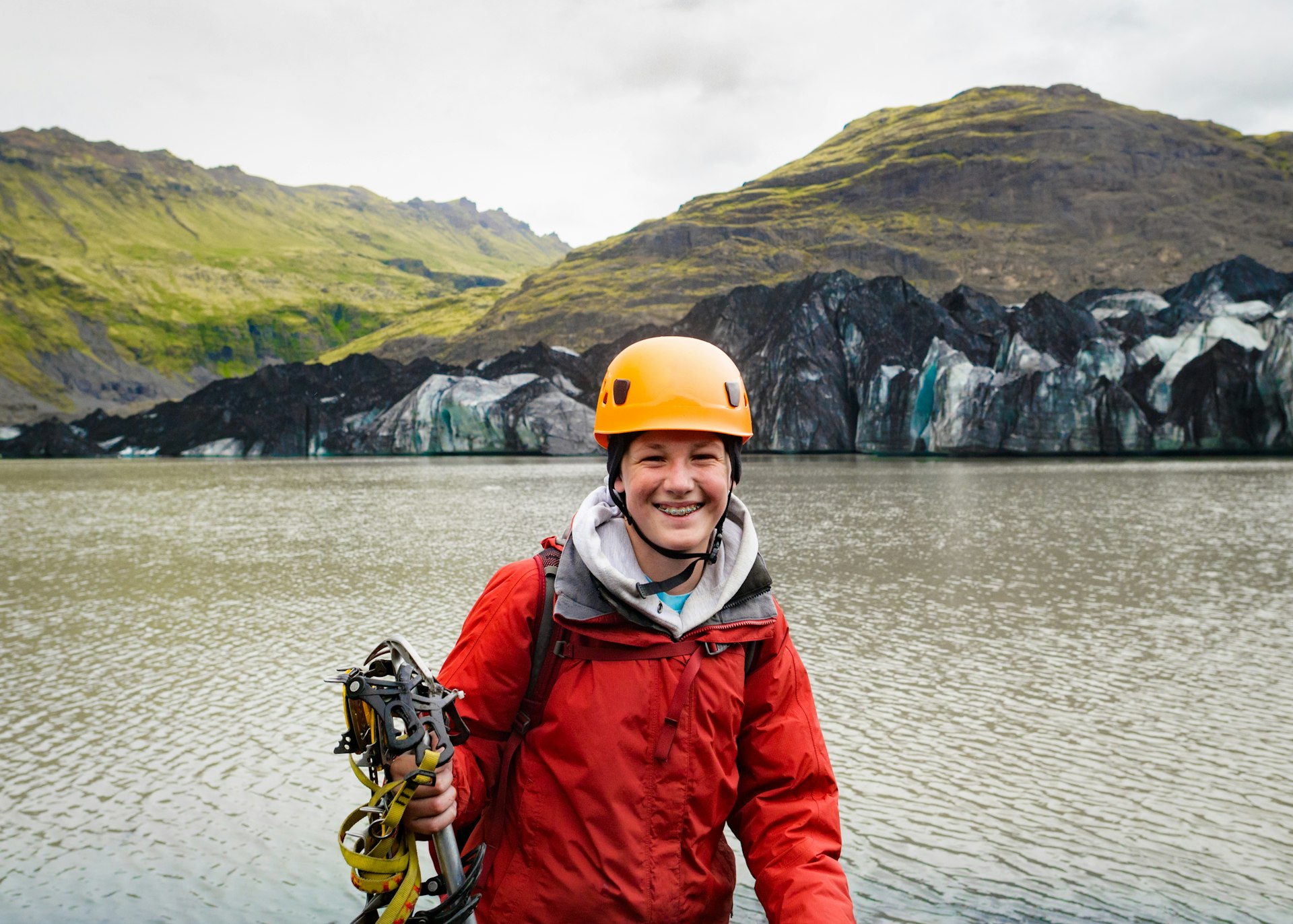Teenage boy wearing ice climbing gear smiling in front of a glacier lake and blue colored ice glacier, Sólheimajökull, Iceland