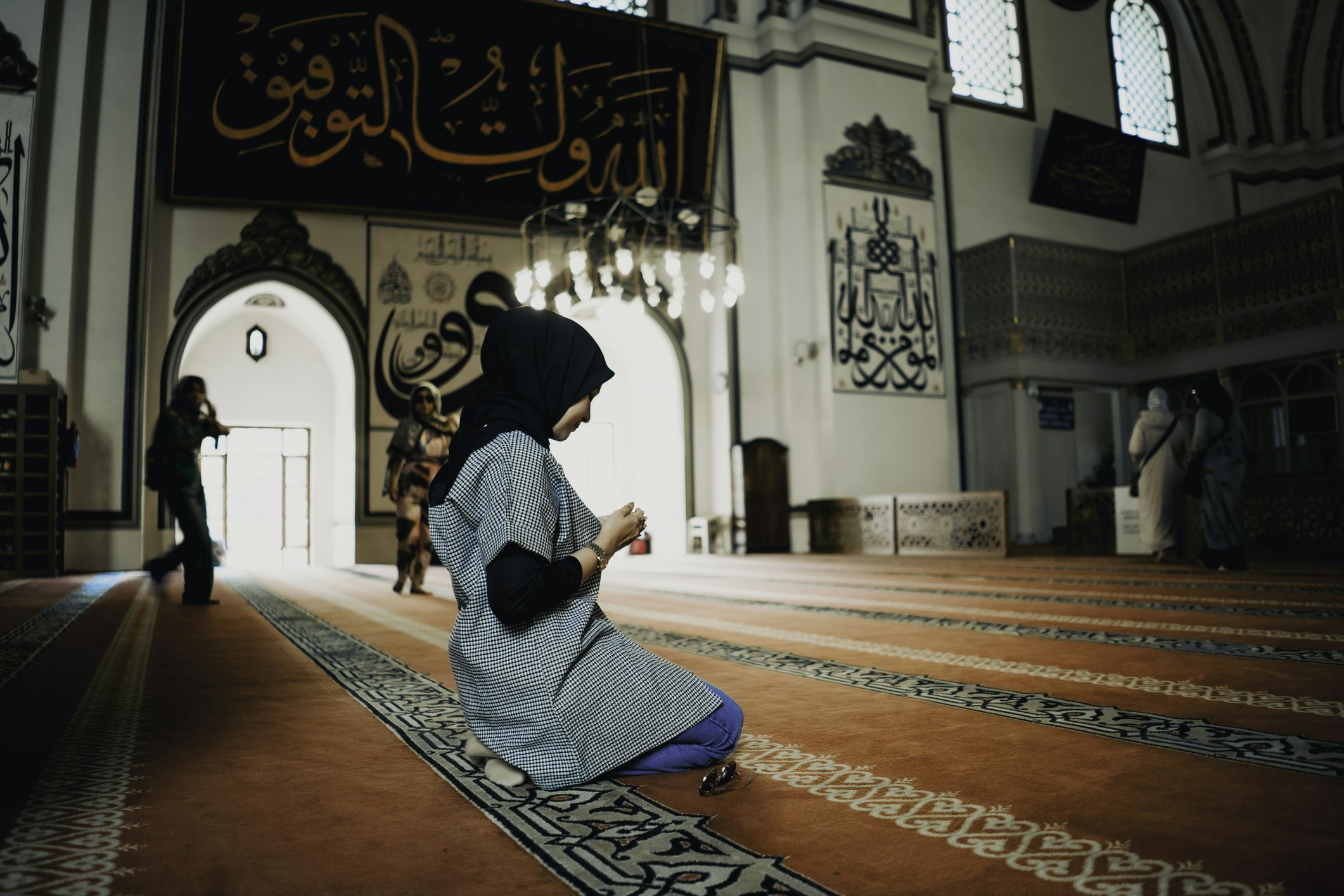 A woman kneels on a carpet in a mosque and holds her hands in front of her in prayer