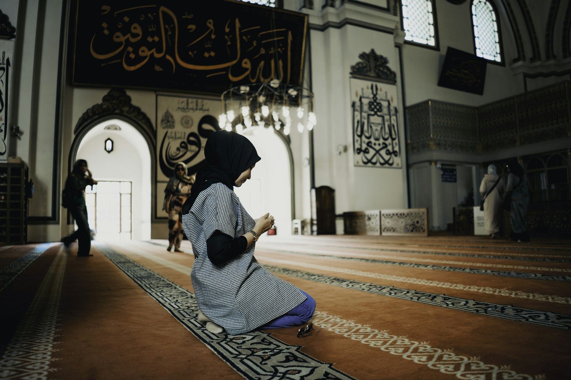 A woman kneels on a carpet in a mosque and holds her hands in front of her in prayer