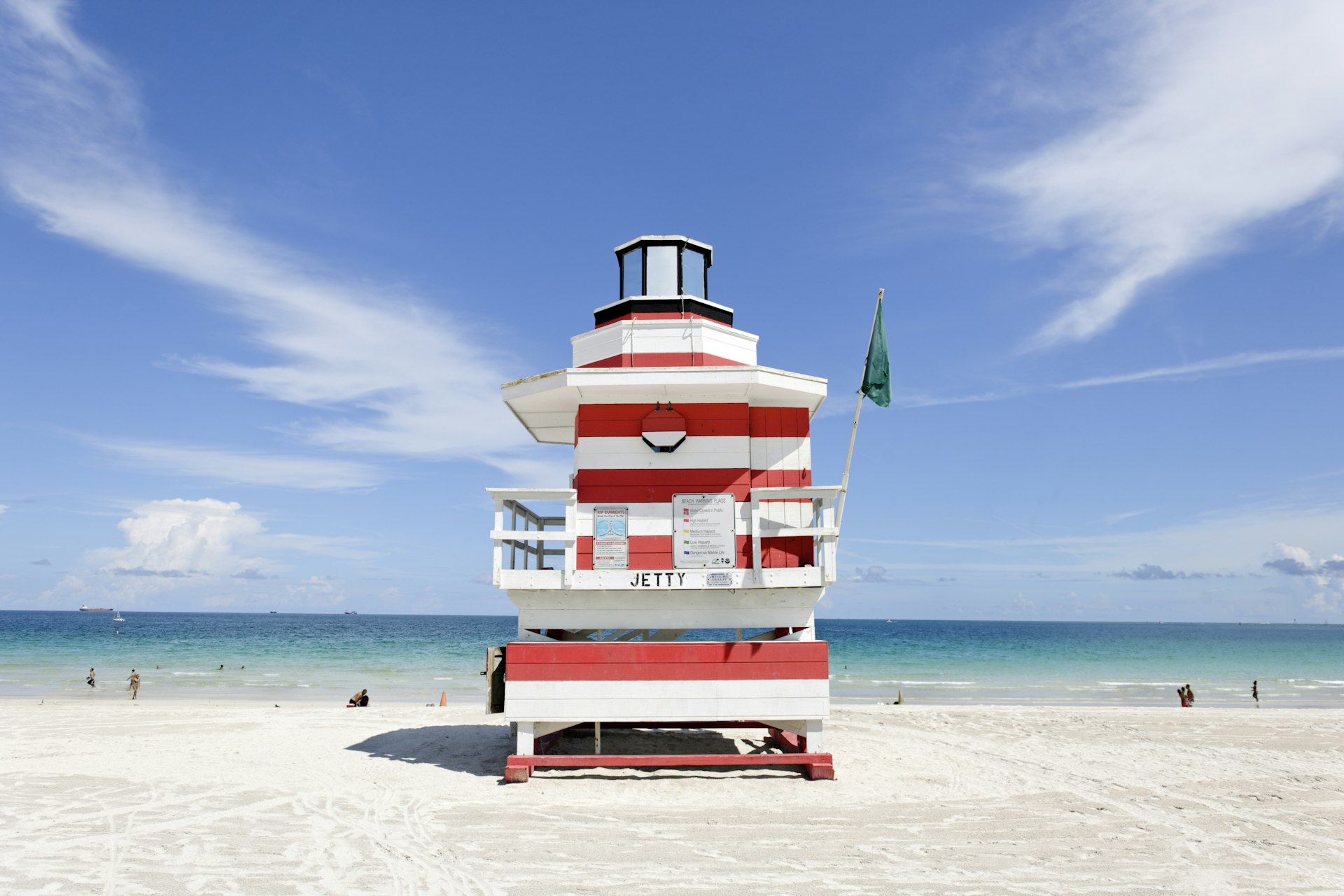 A large red-and-white-striped lifeguard tower standing on a white-sand beach and set against a blue sky with wispy clouds