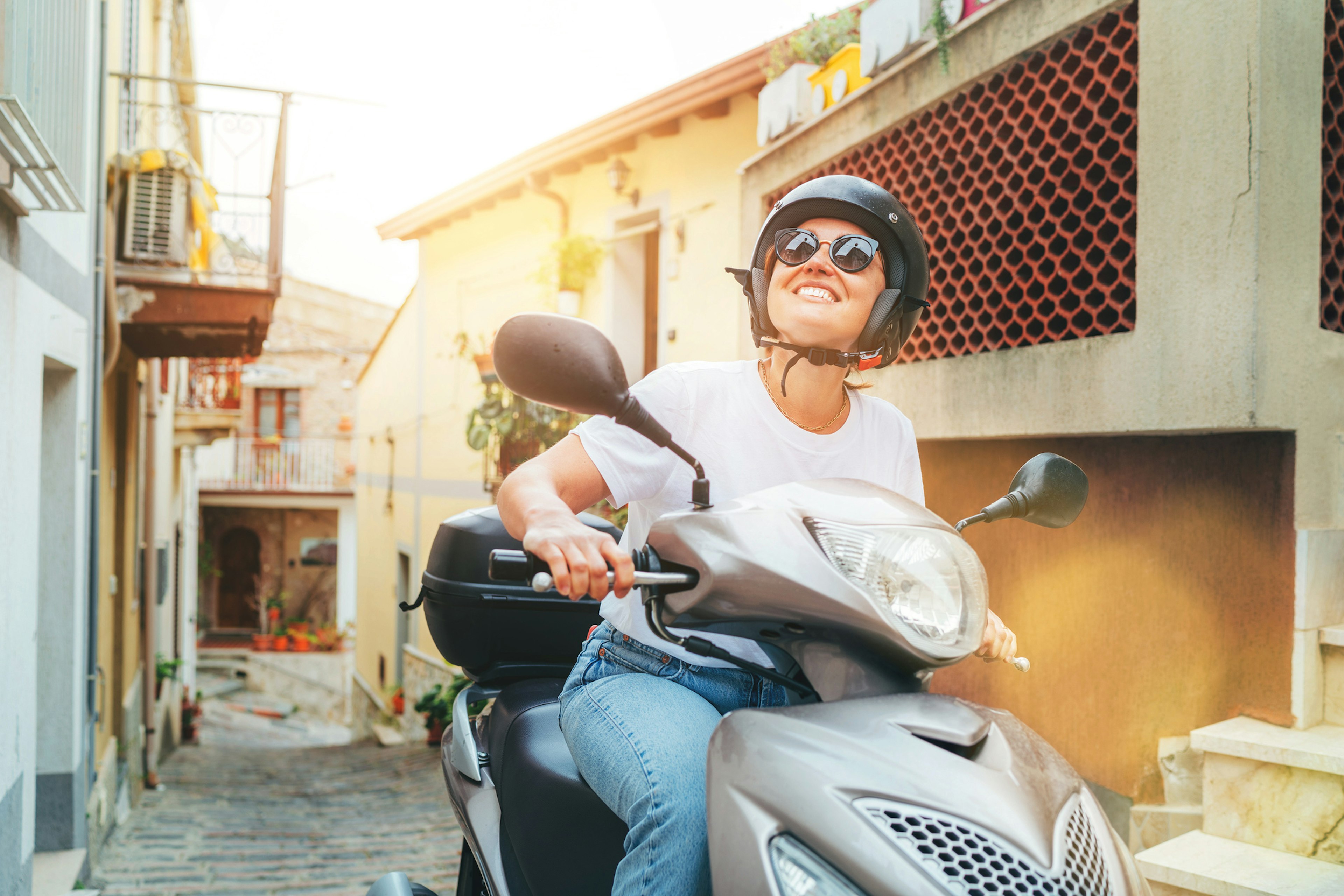 A smiling woman driving a scooter through a narrow street in an Italian town