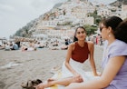 Two pretty young woman sit on a beach towel. They enjoy spending time together, and admire the view. The beach is busy. View of Positano visible behind them.
1494384131
