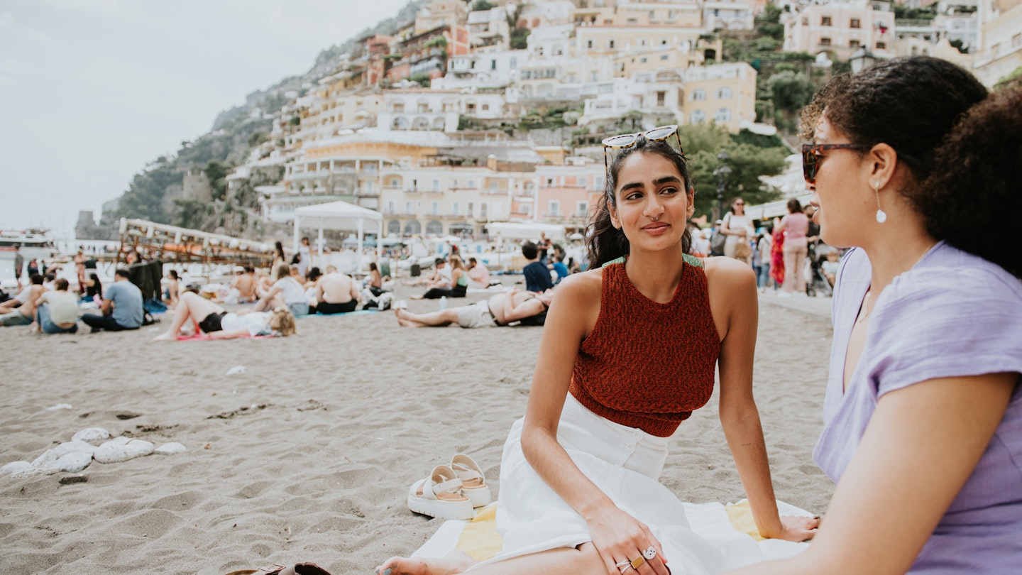 Two pretty young woman sit on a beach towel. They enjoy spending time together, and admire the view. The beach is busy. View of Positano visible behind them.
1494384131