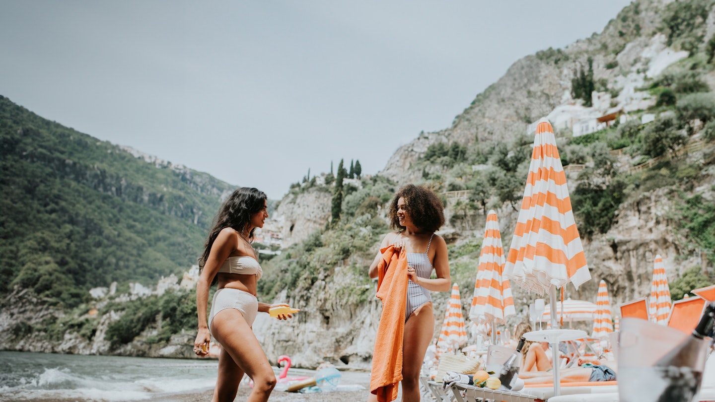 Two beautiful young female friends stand on an idyllic beach in Italy. One female shakes out / folds a large orange beach towel whilst her companion applies sunscreen to her body.
1504361029