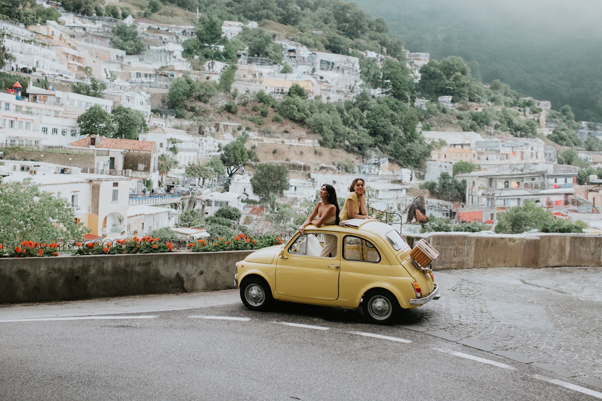 Two woman stand in a vintage yellow fiat, and admire the view of Positano, Italy
