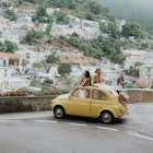 Two woman stand in a vintage yellow fiat, and admire the view of Positano, Italy