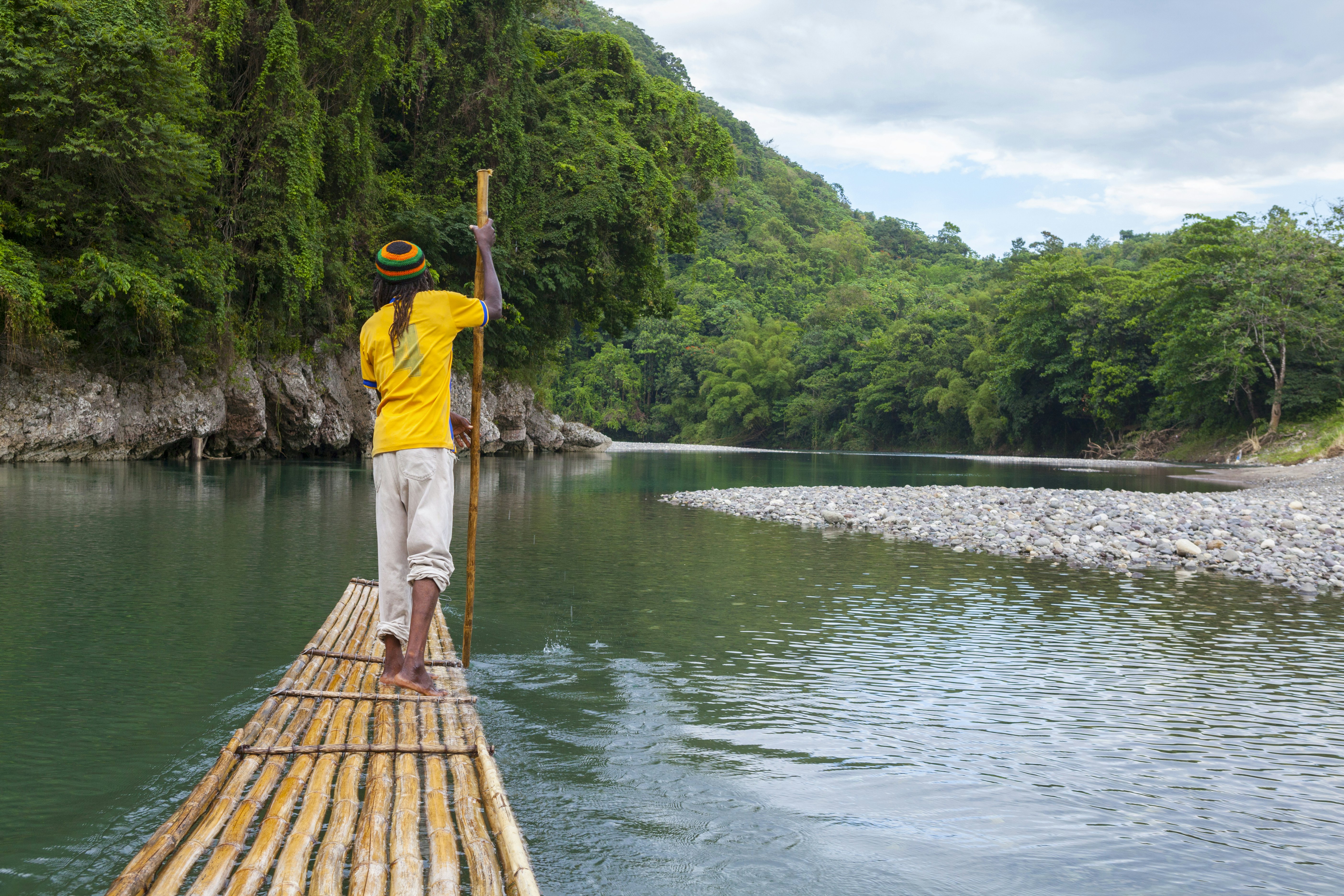 A man in a yellow shirt and striped hat stands at the front of a bamboo raft, pushing it forward through water with a long pole in the water; the waterway is lined by lush green vegetation, and a rocky shore is to the right.