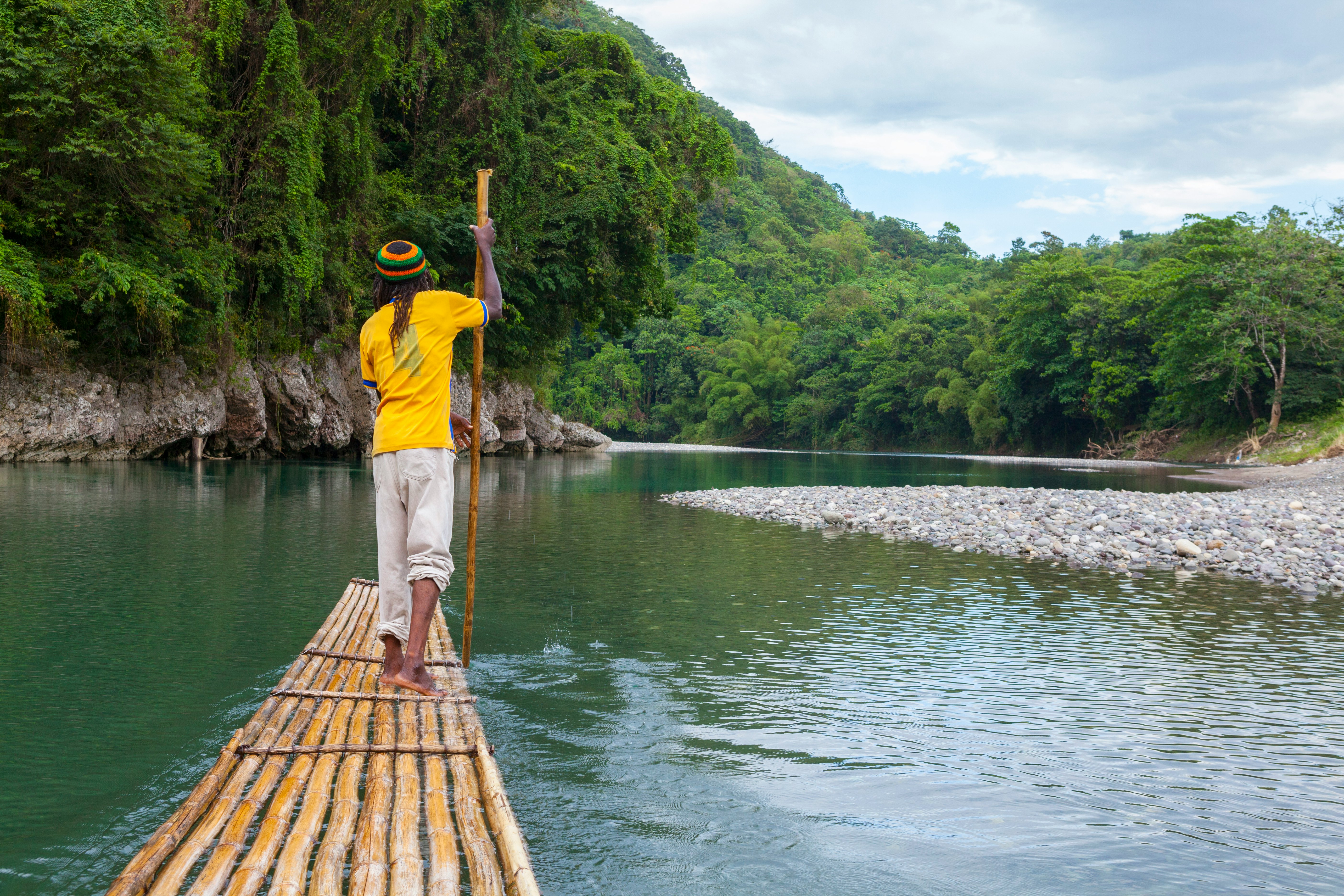 A man in a yellow shirt and striped hat stands at the front of a bamboo raft, pushing it forward through water with a long pole in the water; the waterway is lined by lush green vegetation, and a rocky shore is to the right.