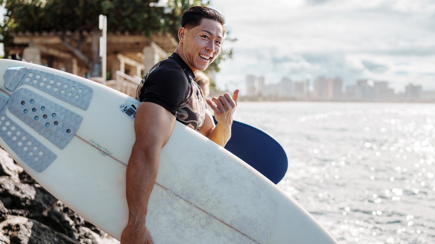 Portrait of a young and fit male surfer of Hawaiian and Japanese descent smiling and holding the shaka sign while walking on a rocky beach with his surfboard with Honolulu, Hawaii visible in the background.
1898786282