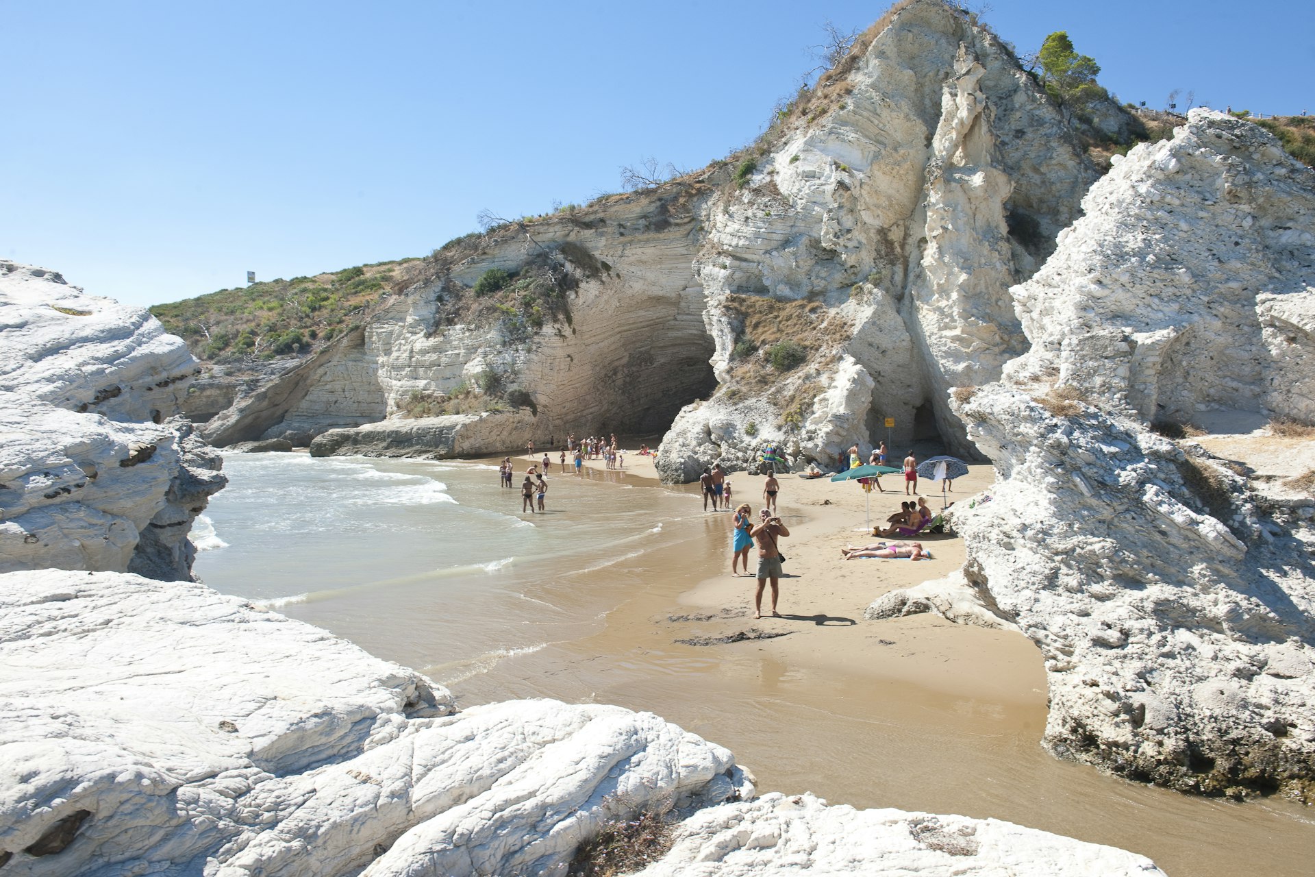 Pessoas caminham entre formações rochosas brancas na praia de Pizzomunno, perto de Vieste