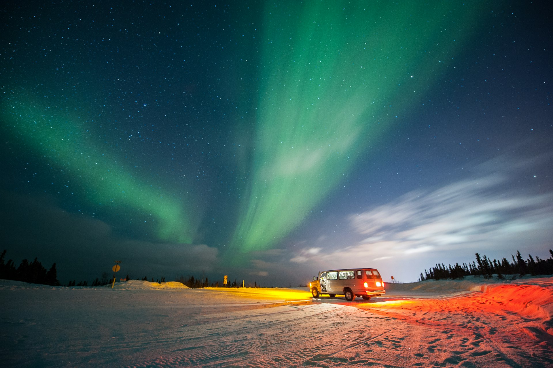 A van is parked with its headlights on, casting an orange glow on the snowy ground, while the aurora borealis lights up the sky with vibrant streaks of green