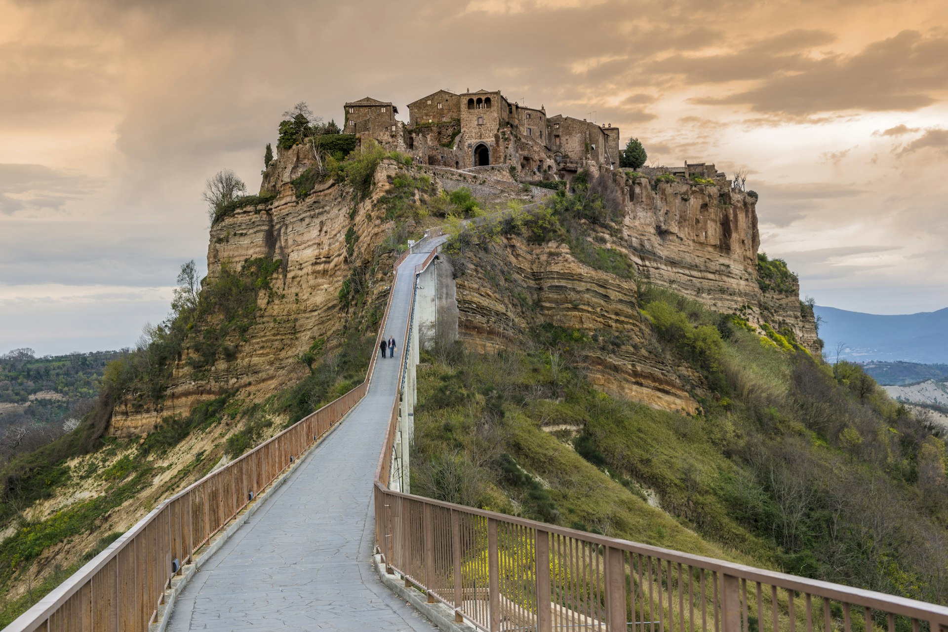 A hilltop crowned with a medieval town. Two people walk down the narrow path that leads down from the hill