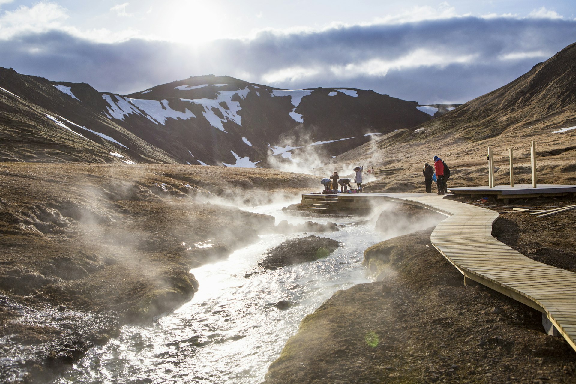 People walk on boardwalks alongside hot springs with steam rising from the water into the cold air