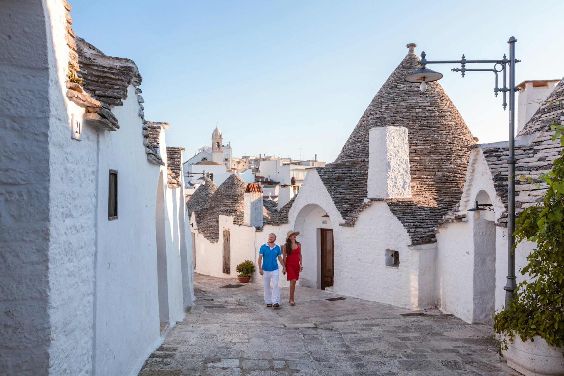 A couple walks past dome-shaped trulli houses on a street in Alberobello, Valle d’Itria, Puglia, Italy