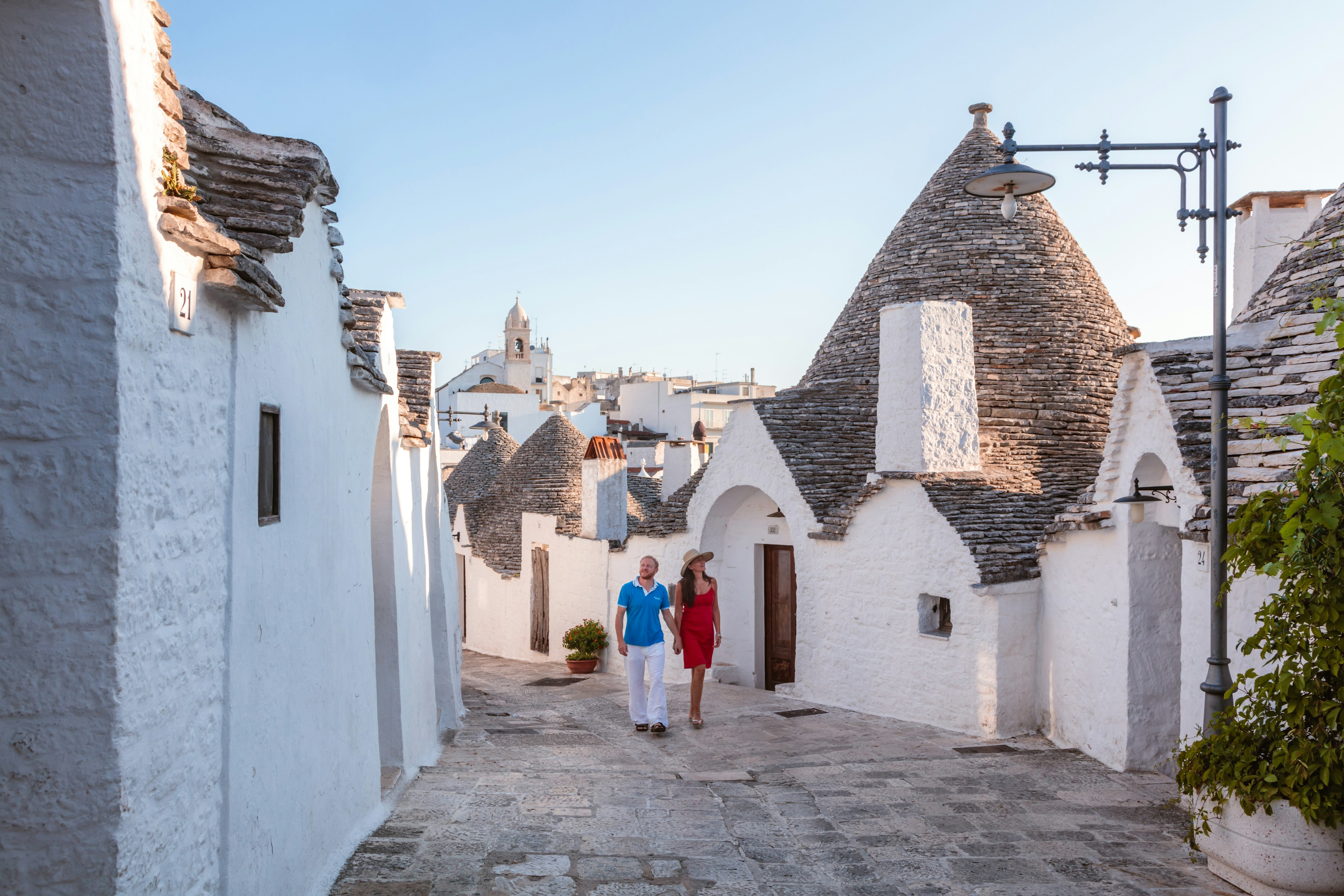 A couple walks past dome-shaped trulli houses on a street in Alberobello, Valle d’Itria, Puglia, Italy