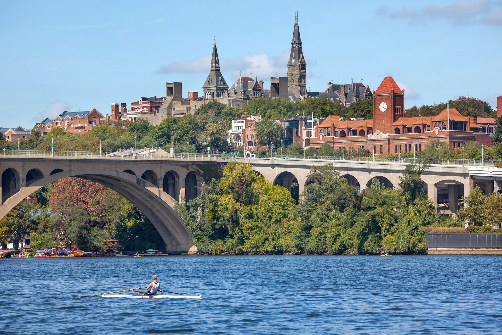 A kayaker on a river heads towards a bridge. A large Gothic-style building rises up on the river bank