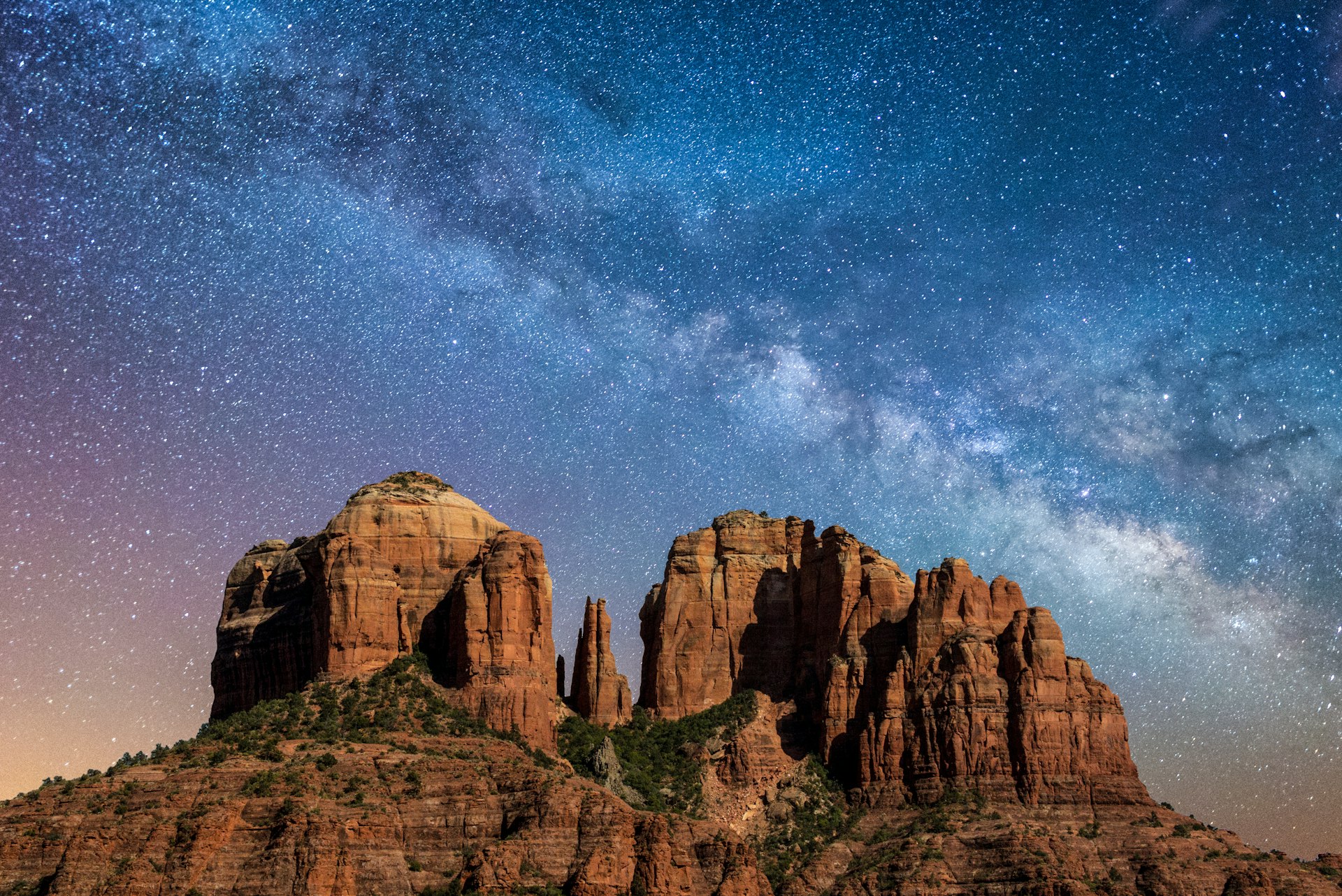 A towering reddish-brown rock formation stands against the night sky, illuminated by the stars of the Milky Way
