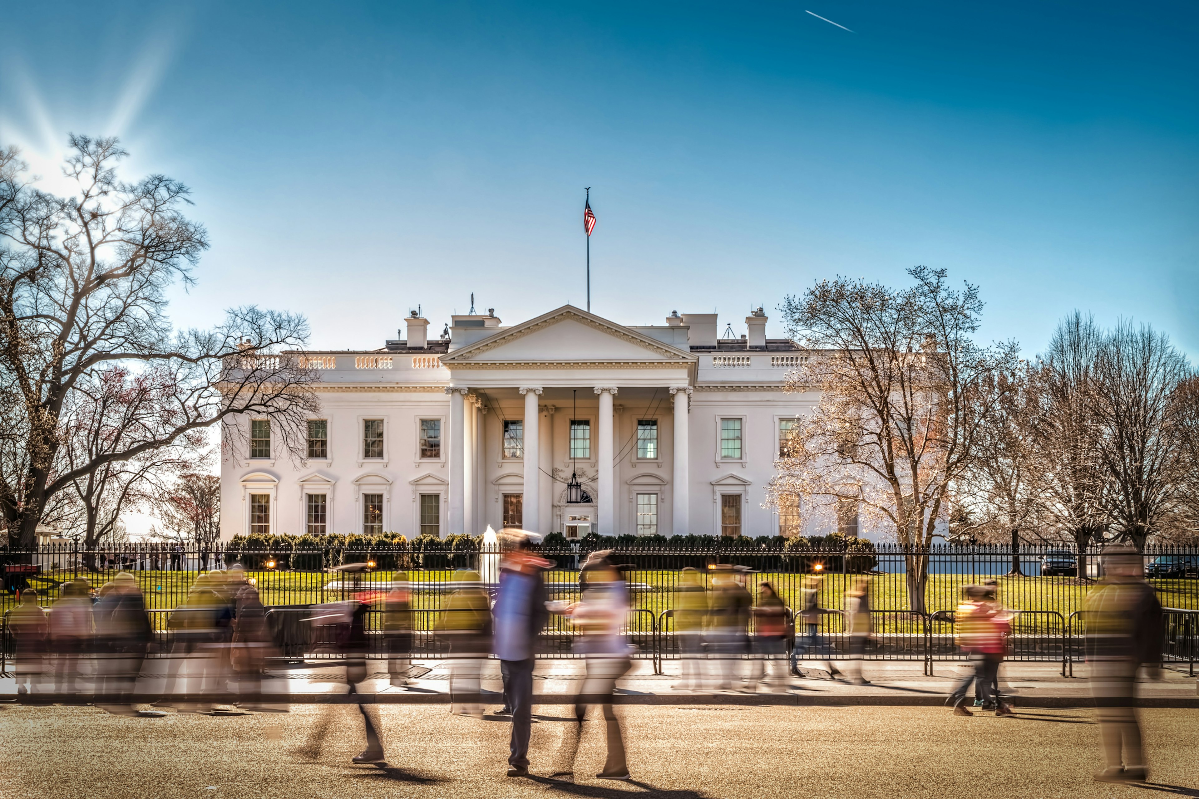 People pass in front of a gated white building set in large grounds with a fountain