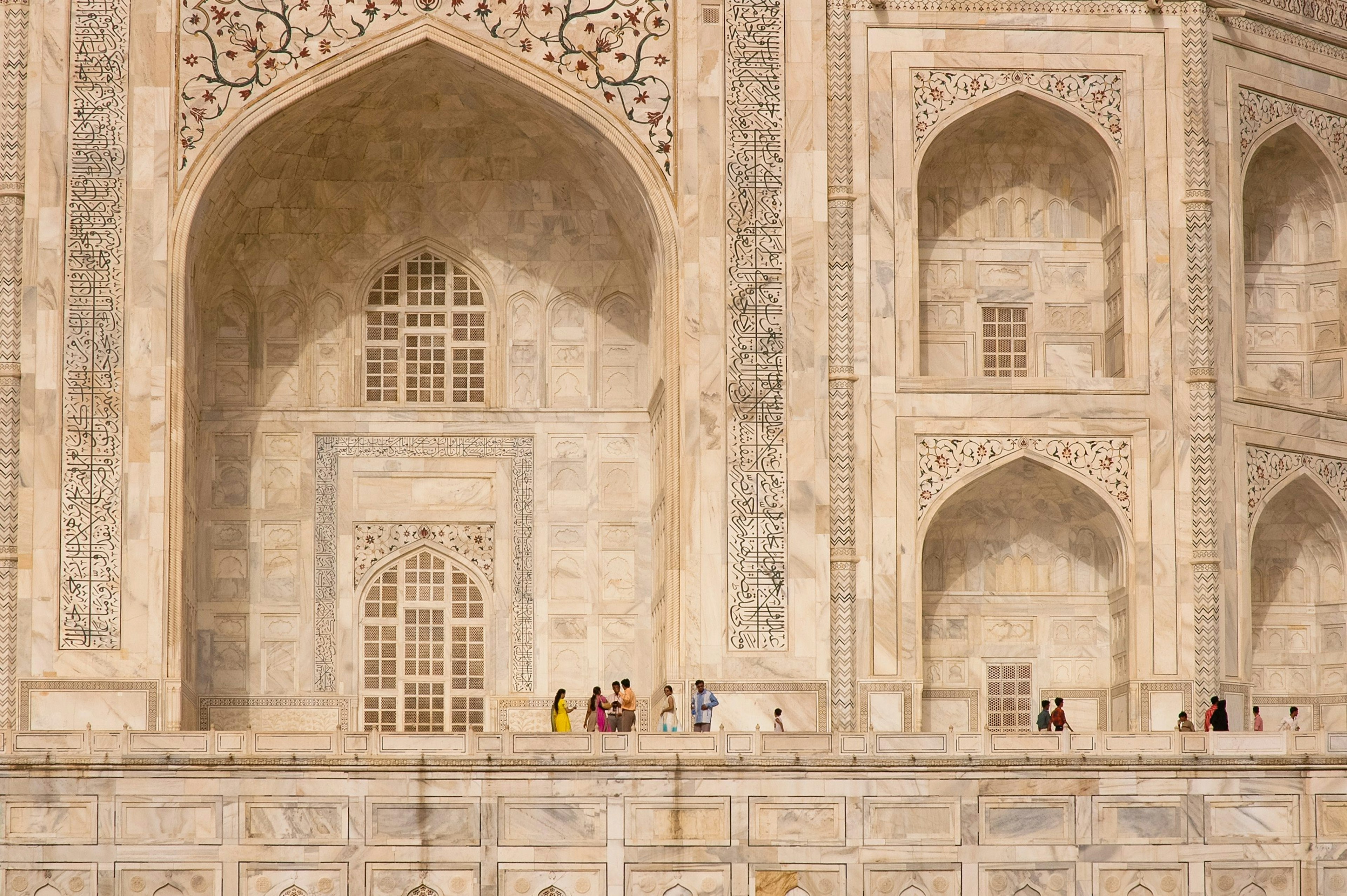 A giant white marble building with a gateway surrounded with intricate calligraphy. People stand at the foot of the building in brightly colored clothes. They look tiny against this giant monument