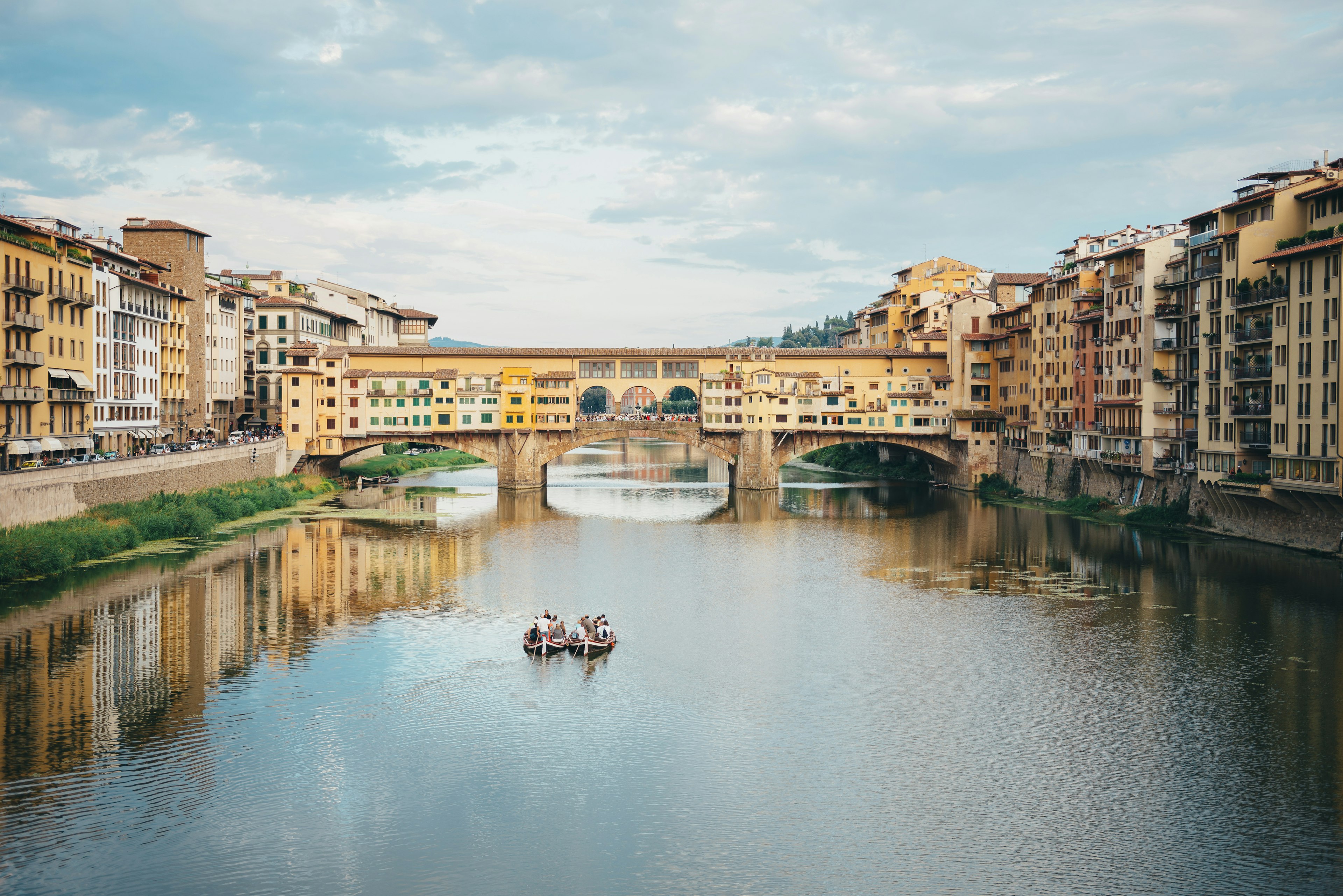 Boaters paddle down a river in a city with a bridge covered in shops up ahead of them