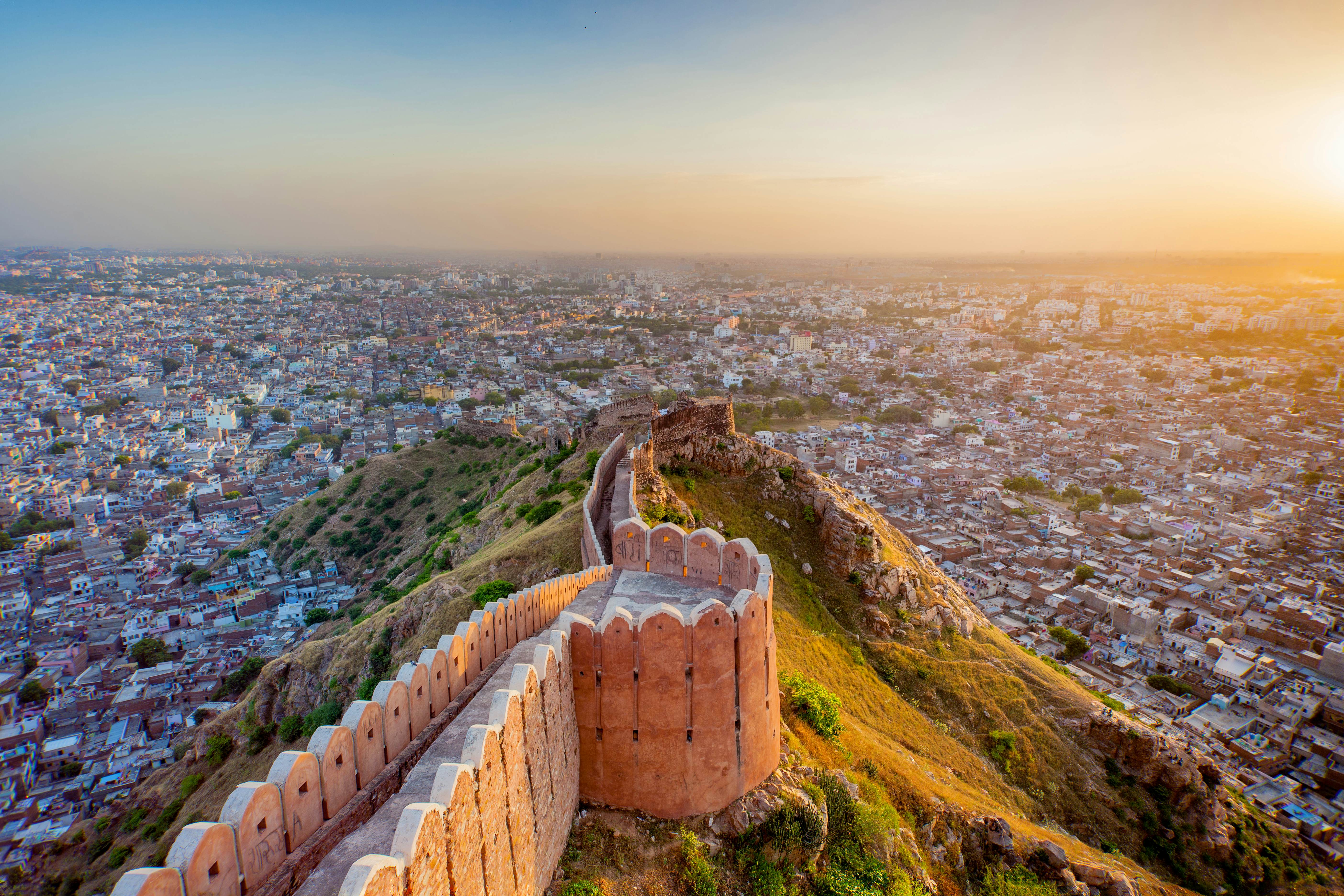 A fortress wall follows the crest of a hill down towards a city glowing in the light of sunset