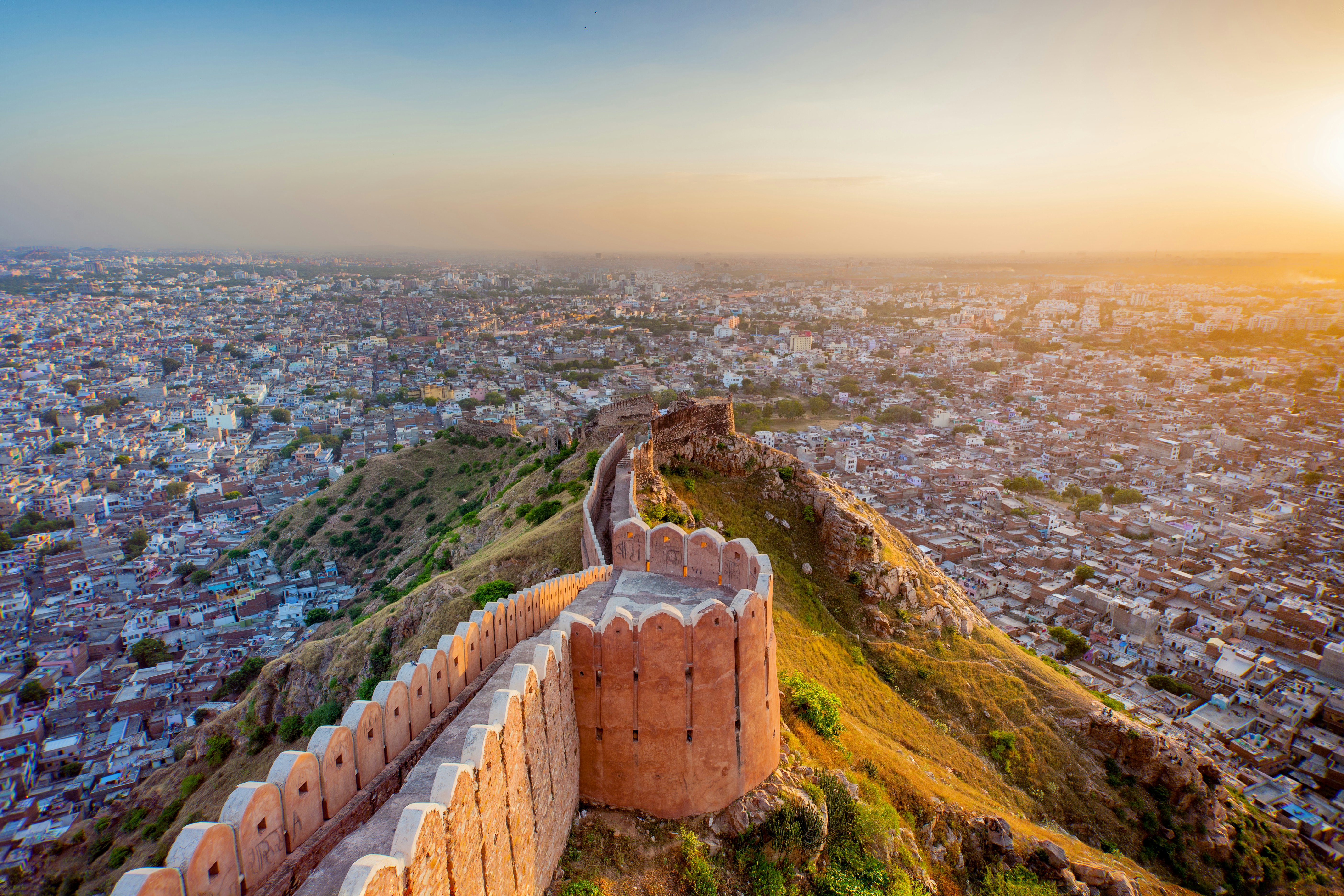 A fortress wall follows the crest of a hill down towards a city glowing in the light of sunset