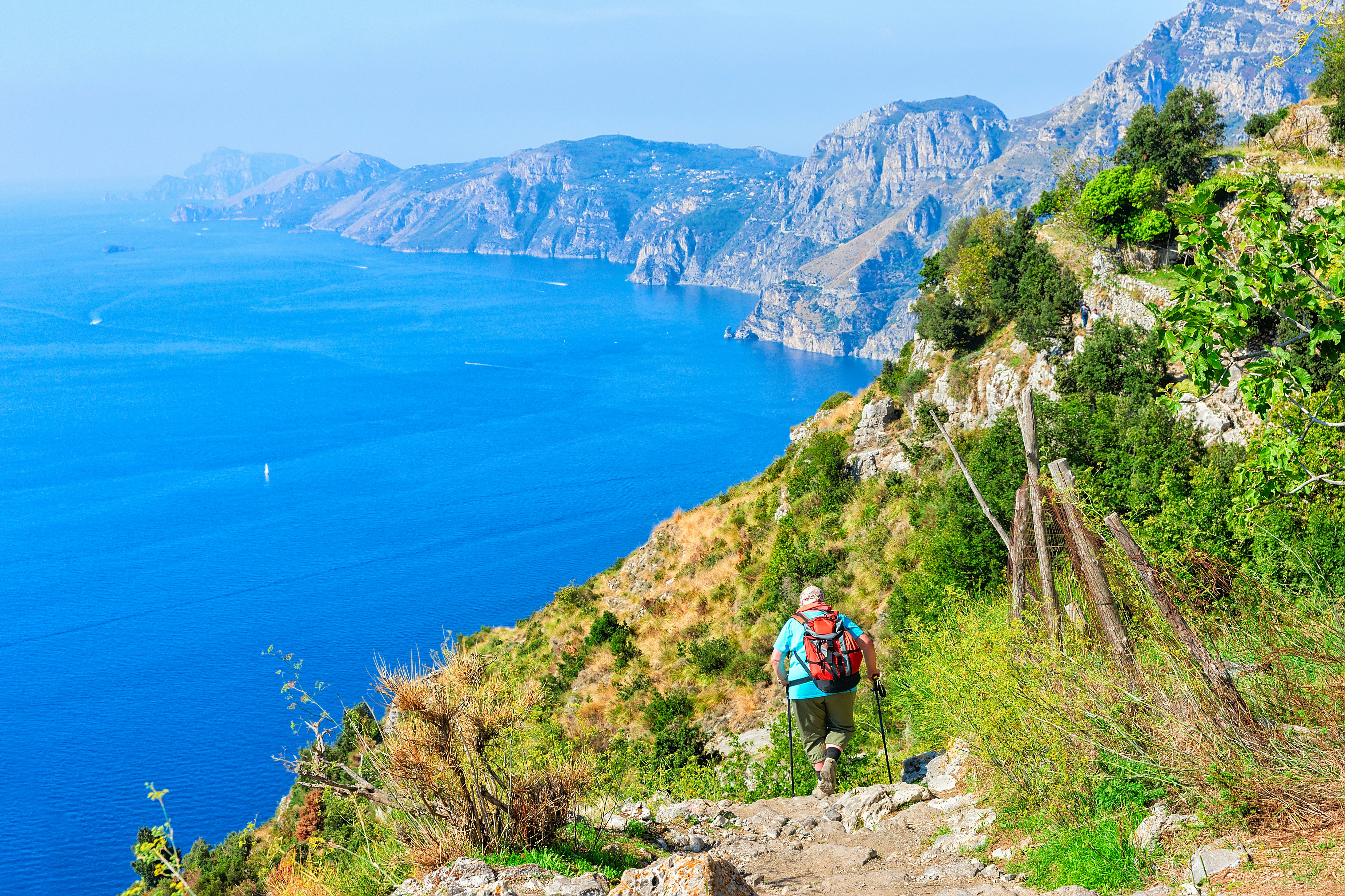 A hiker follows a coastal path with the blue ocean and green hills