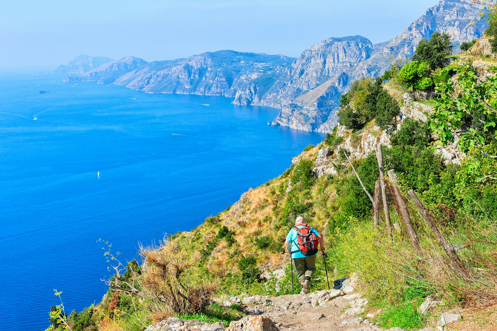 A hiker follows a coastal path with the blue ocean and green hills