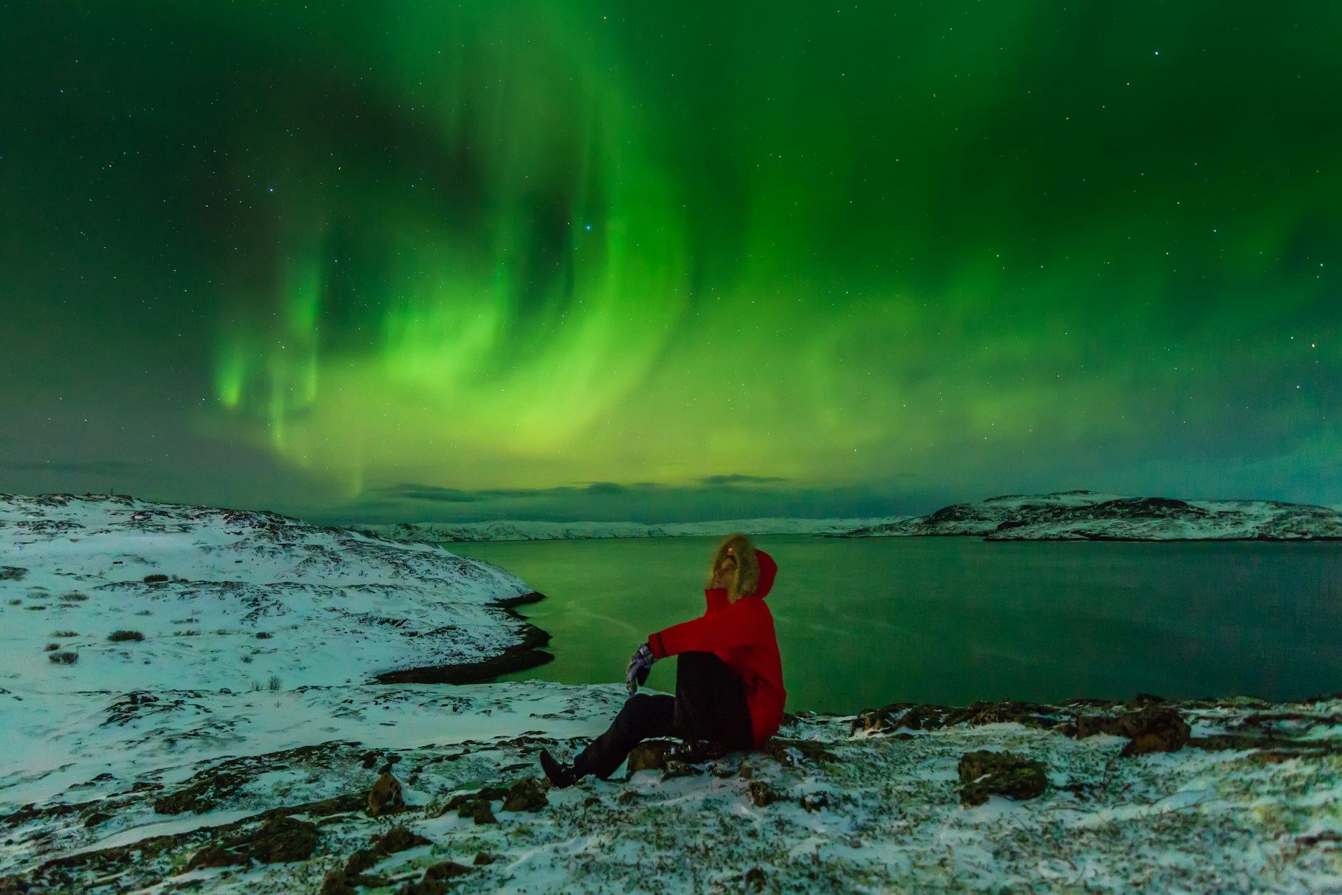 A man sits on the snow with the northern lights in the sky on the North Sea shore, Iceland