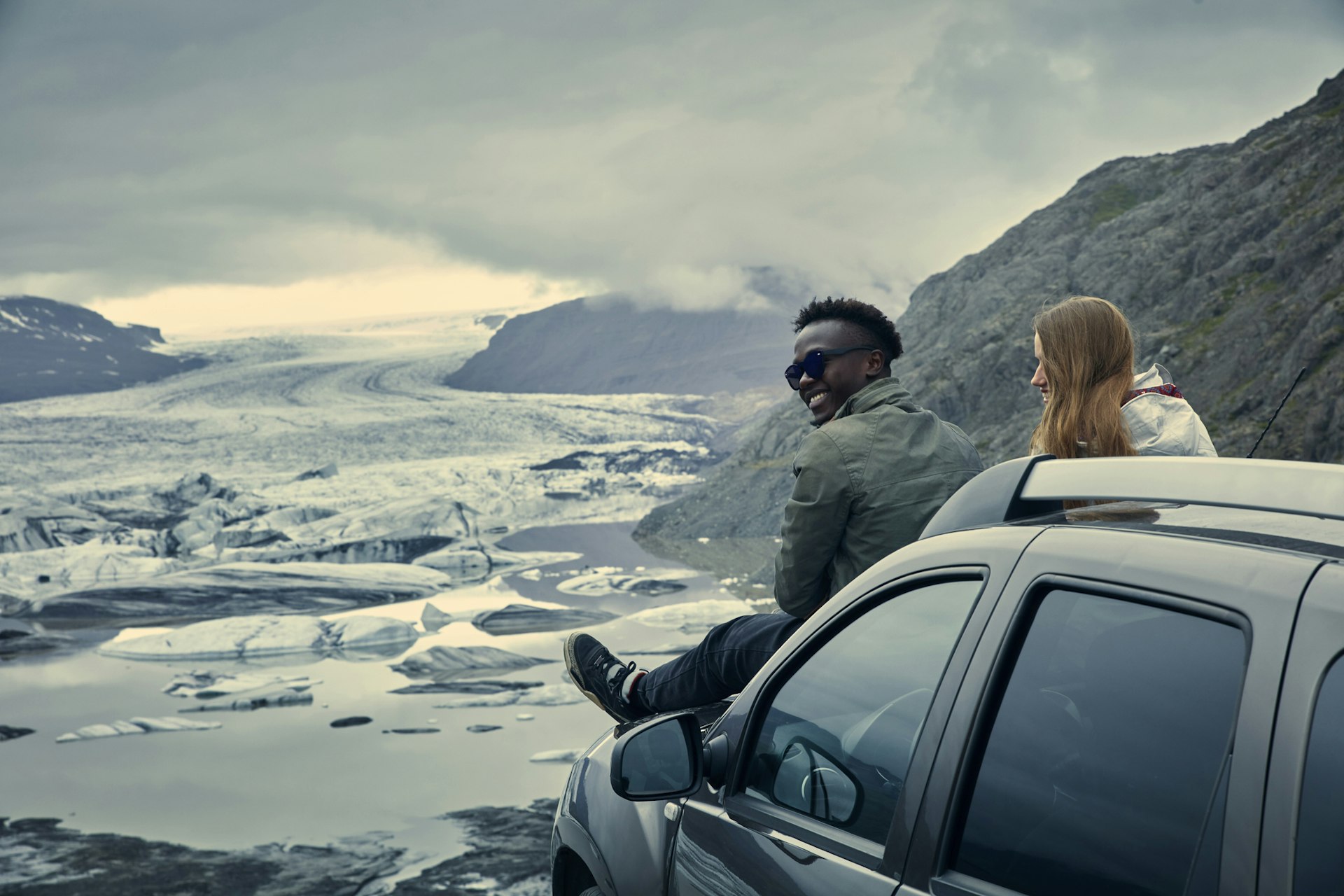 A couple sits on the hood of their car as they look out at a glacier in Iceland
