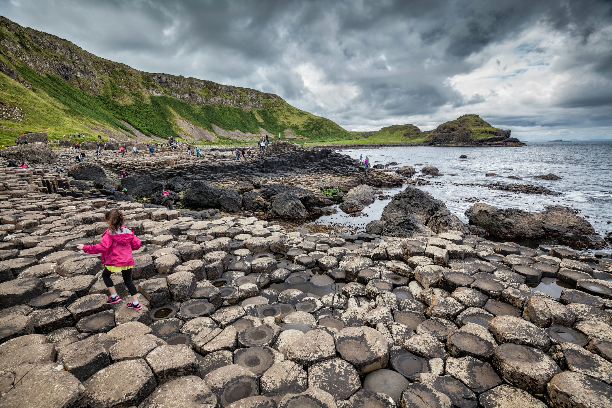 A little girl wearing a pink jacket is walking over the basalt columns of the Giants Causeway in Northern Ireland