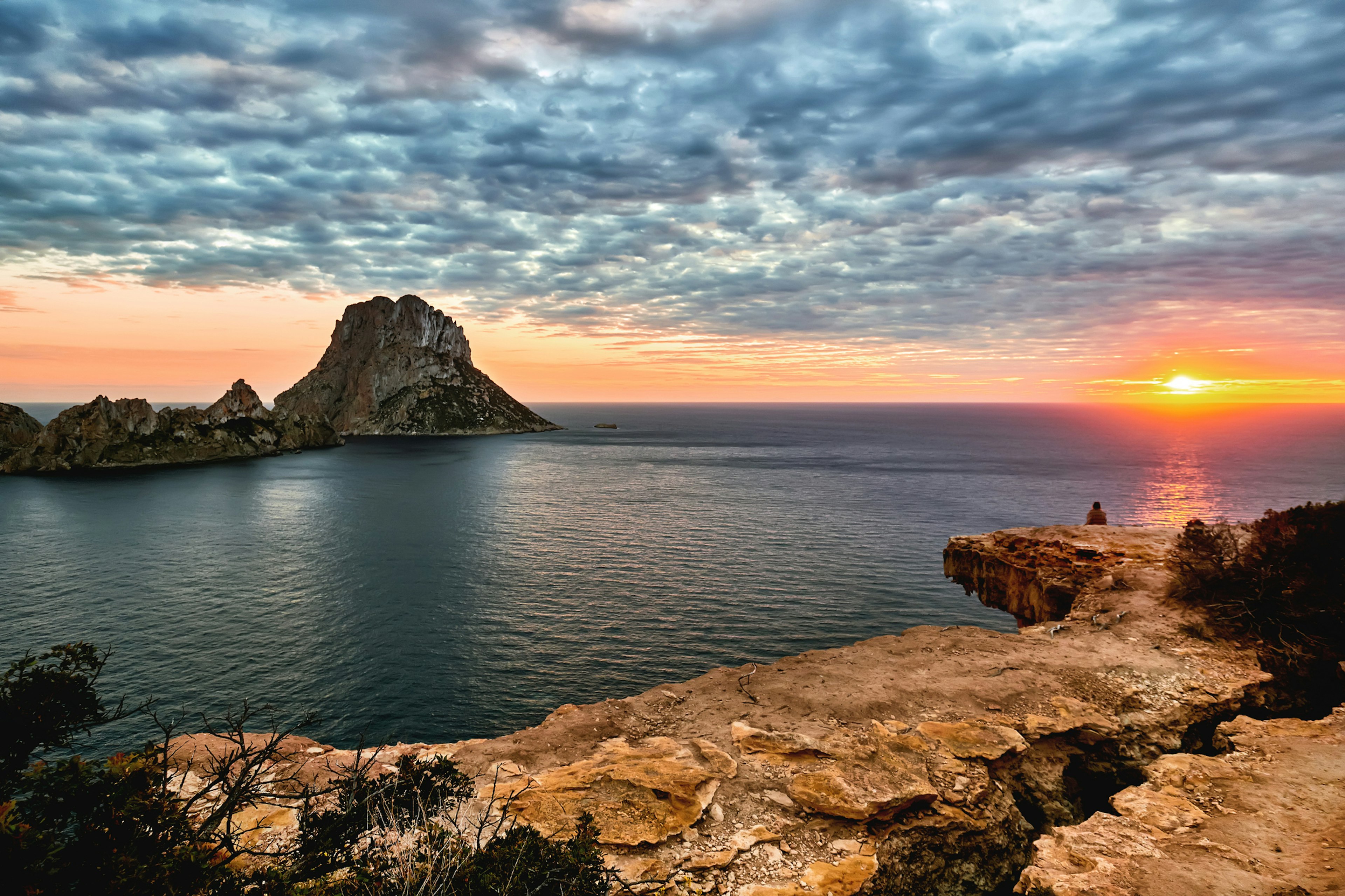 The sun sets over the sea casting an orange glow contrasted by dark grey clusters of cloud. A huge rocky islet stands off shore