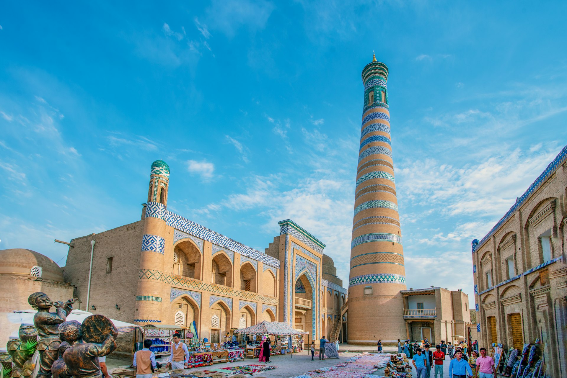 A market selling colorful textiles sets up at the foot of a tall minaret