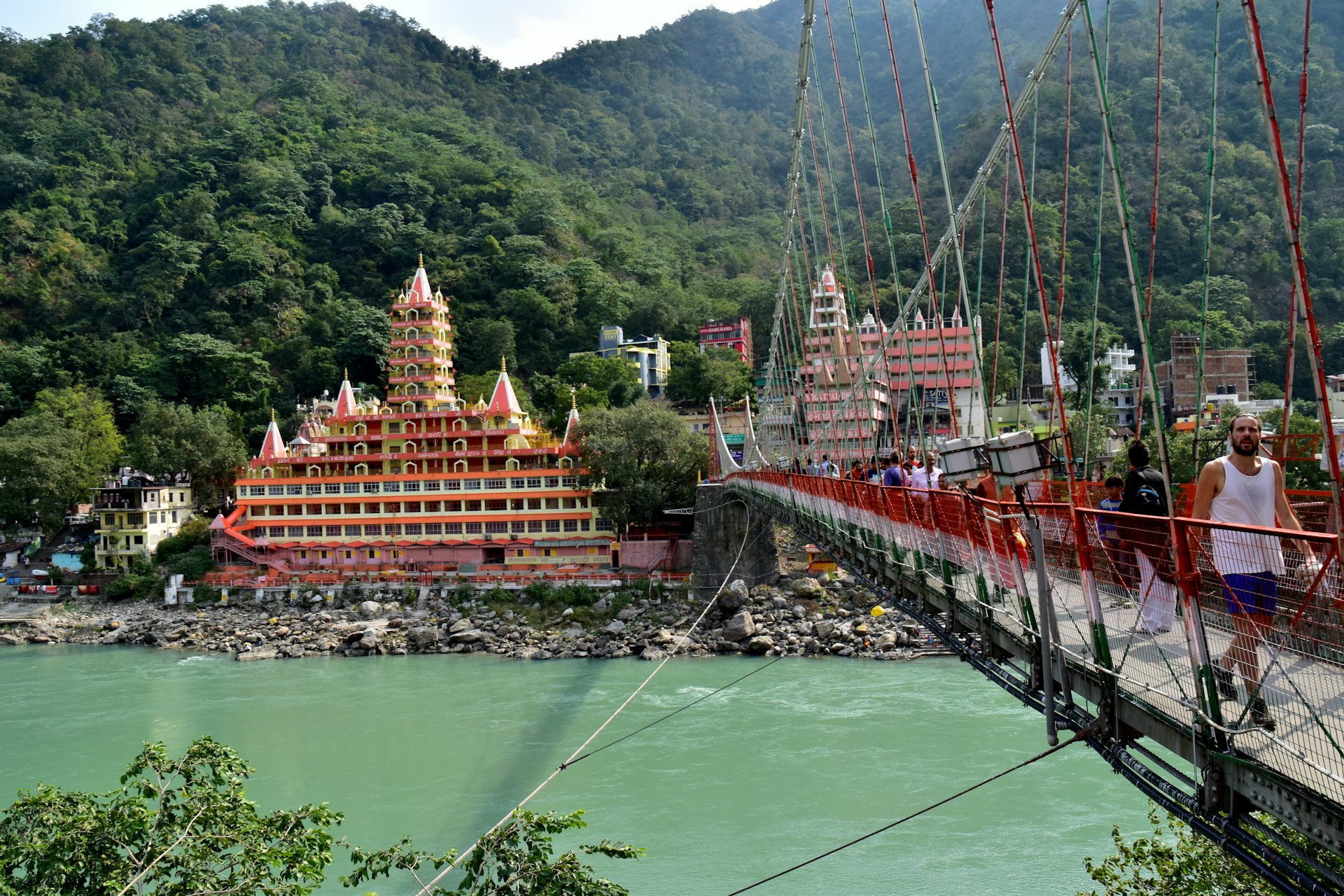 People cross a suspension bridge leading over a wide river towards a temple building surrounded by woodland