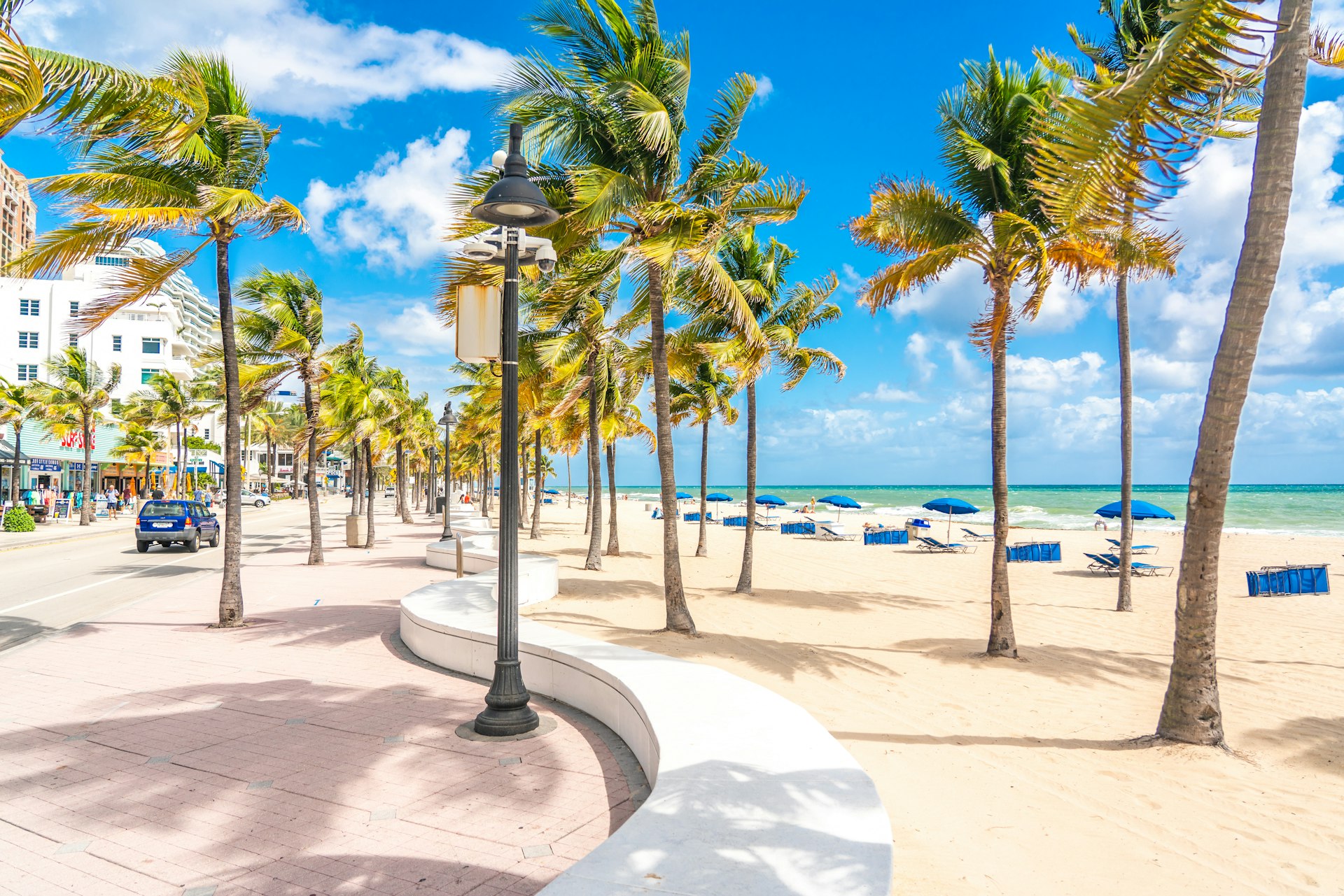 The seafront beach promenade in Fort Lauderdale is lined with palm trees, while blue sun loungers are laid out on the white-sand beach beyond. 