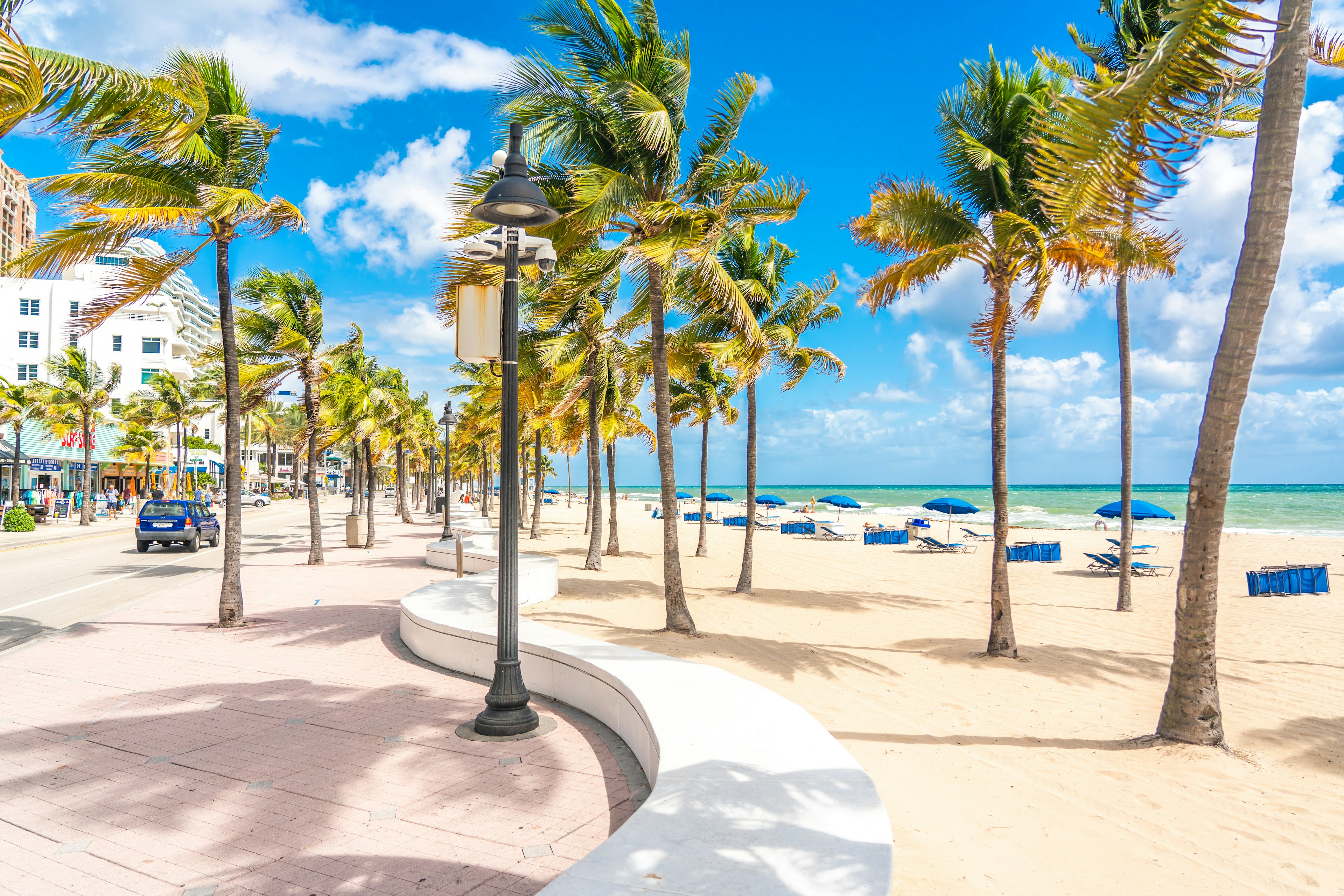 The seafront beach promenade in Fort Lauderdale is lined with palm trees, while blue sun loungers are laid out on the white-sand beach beyond.