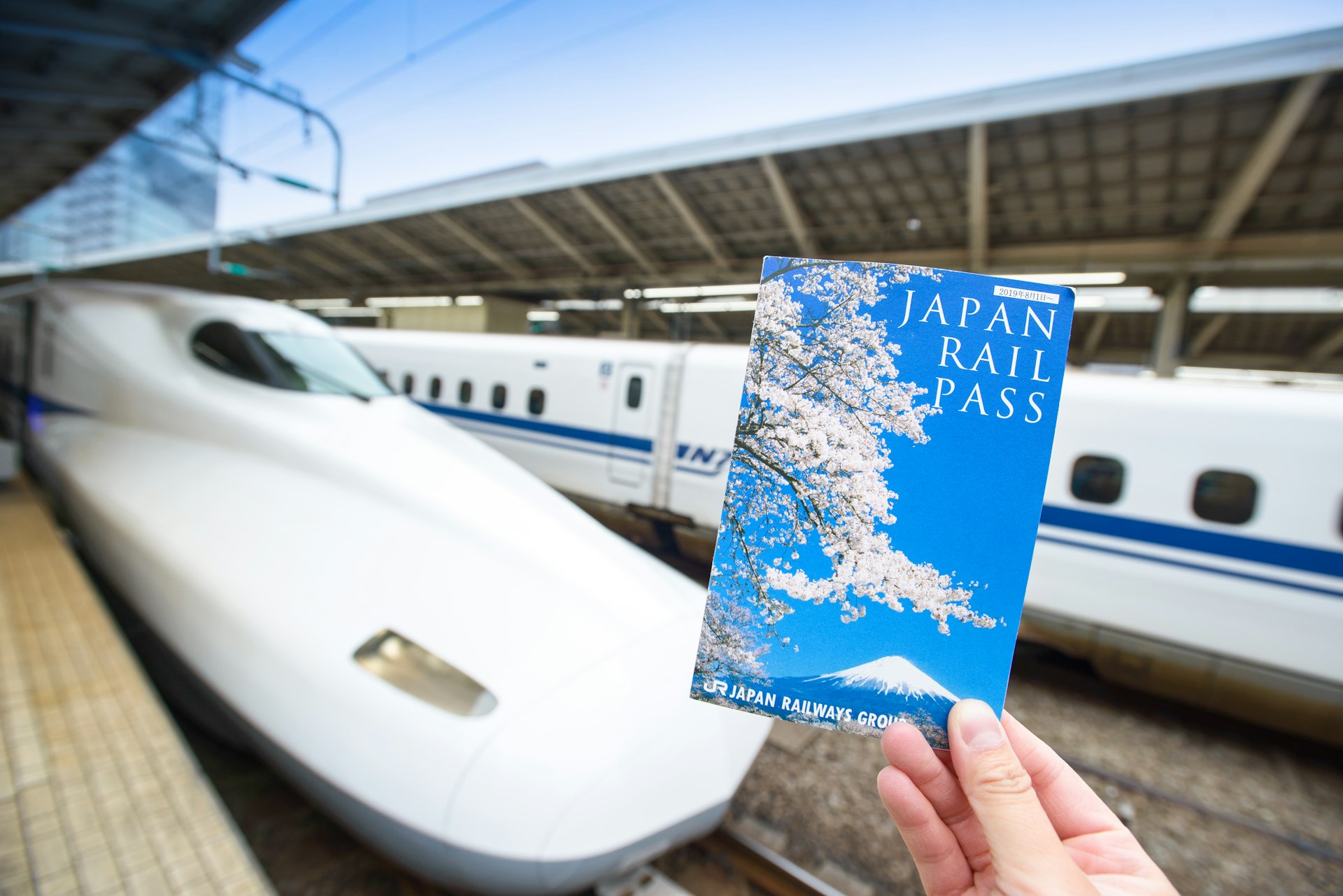 A hand holds up a Japan Rail Pass in front of the rounded nose of a bullet train at a station 