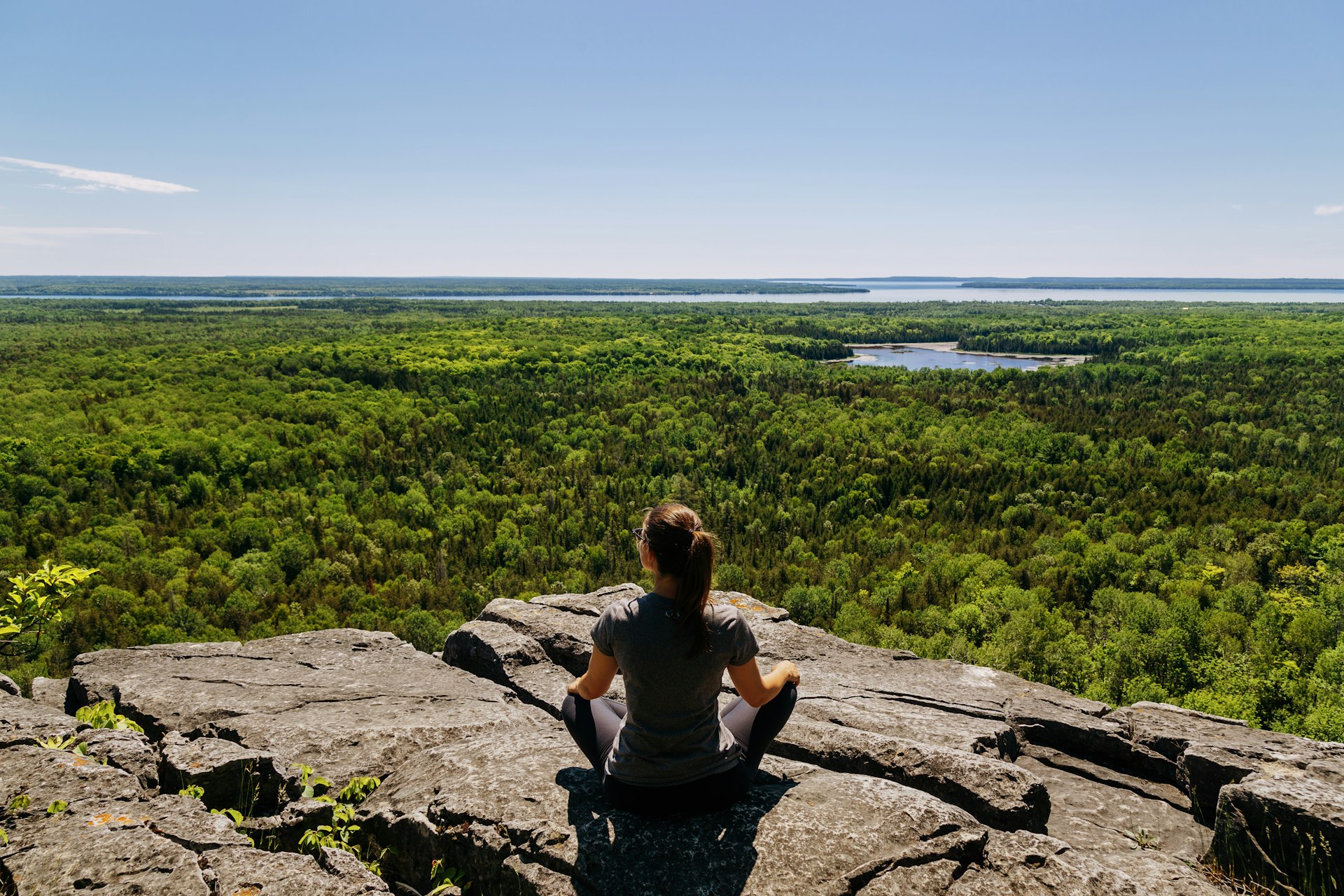 Uma mulher senta-se no topo de uma pedra e olha para a floresta à sua frente em Michigiwadinong (Trilha da Copa e do Pires), Ilha Manitoulin, Ontário, Canadá