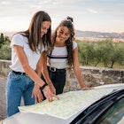 Two women smile and look at a map resting on their car in Italy