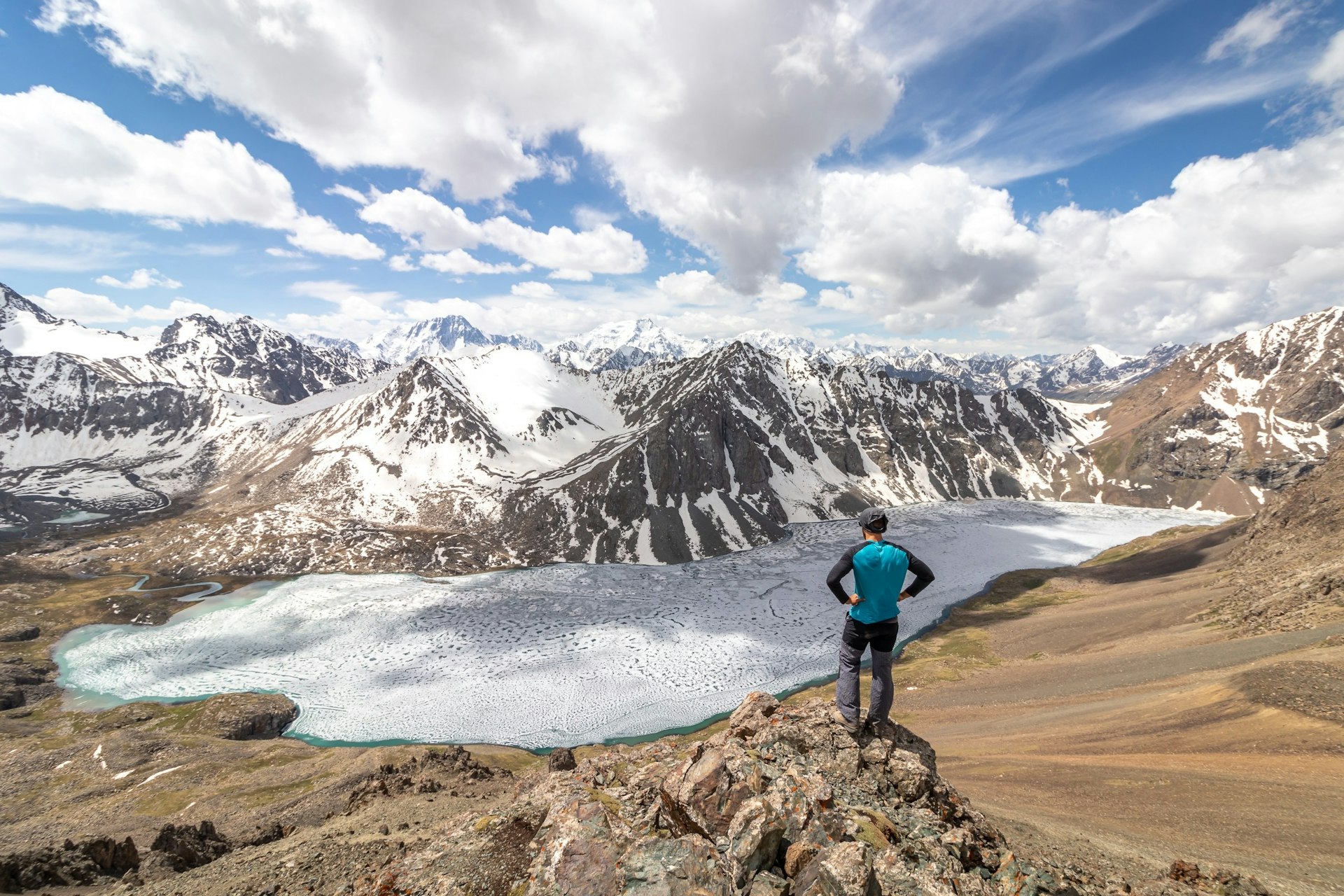 A person stands on the edge of a precipice with a frozen lake below them and a mountain range stretching out before them