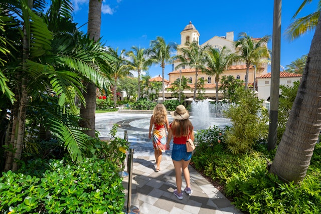 Two women walking on a path through palm trees at The Square shopping complex in West Palm Beach, Florida, USA.