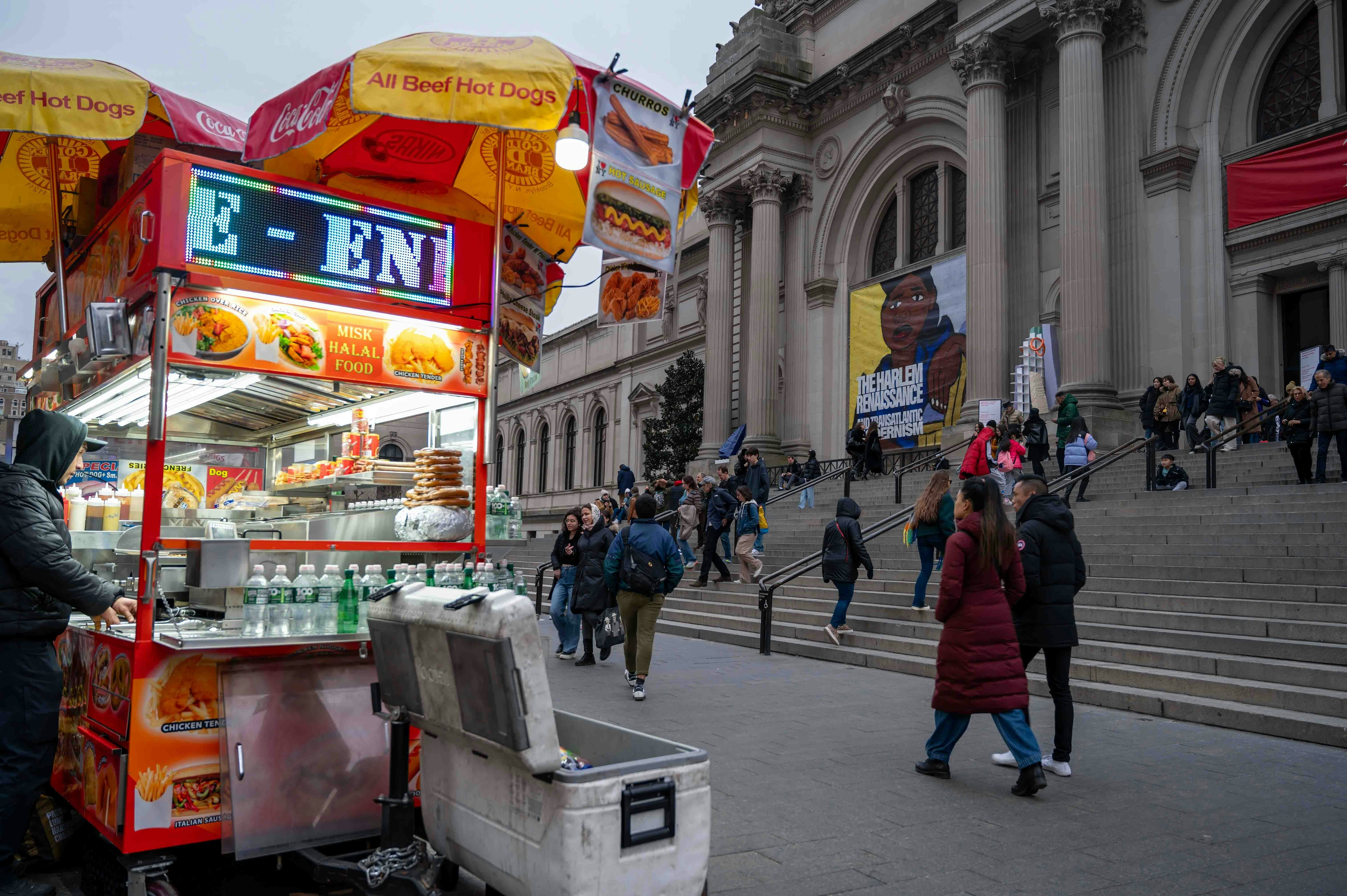 A hot dog stand at the foot of some stairs leading into a museum building on a cold gray day