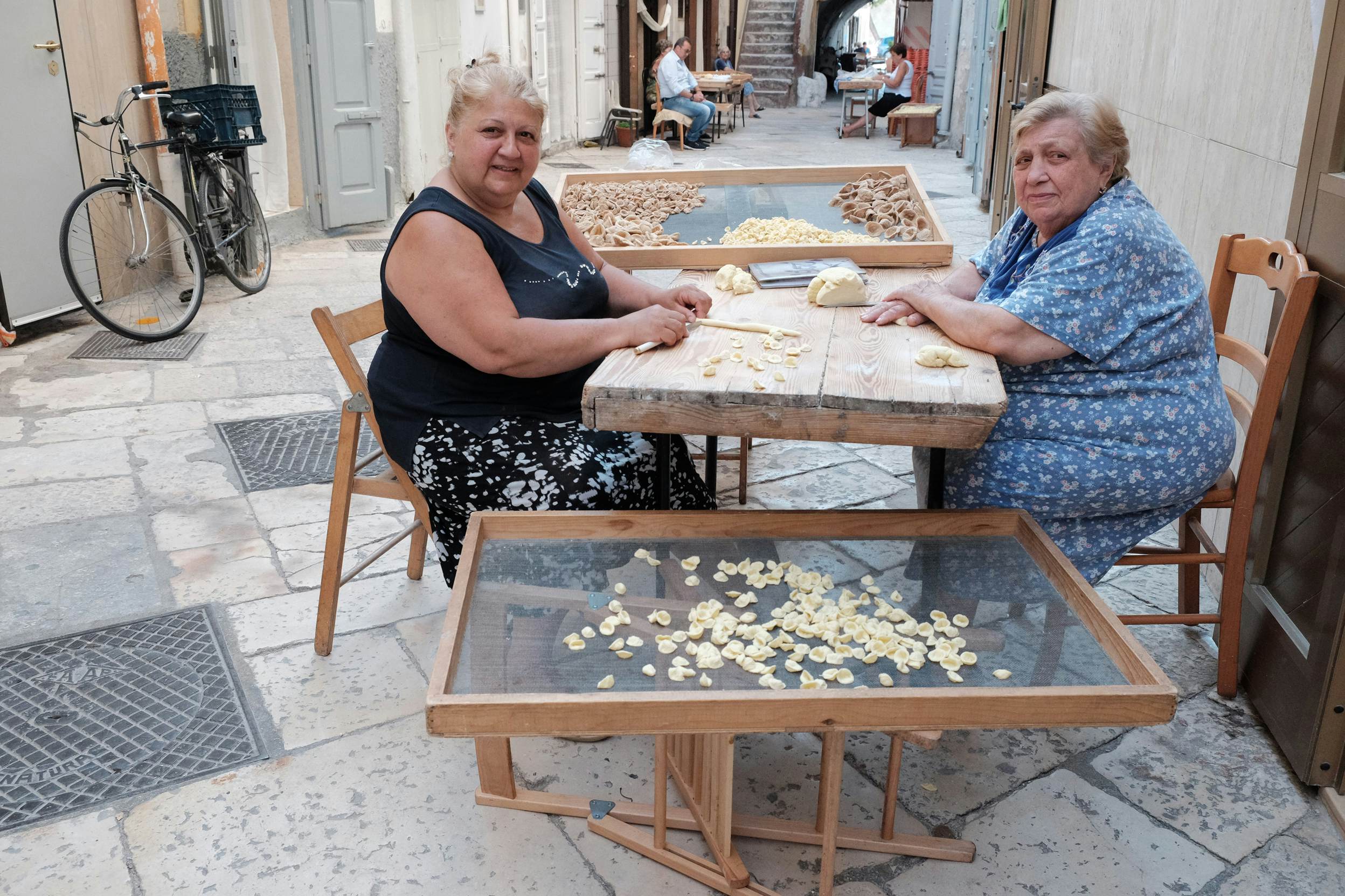 Two women sit at a table on a narrow street rolling pasta by hand.