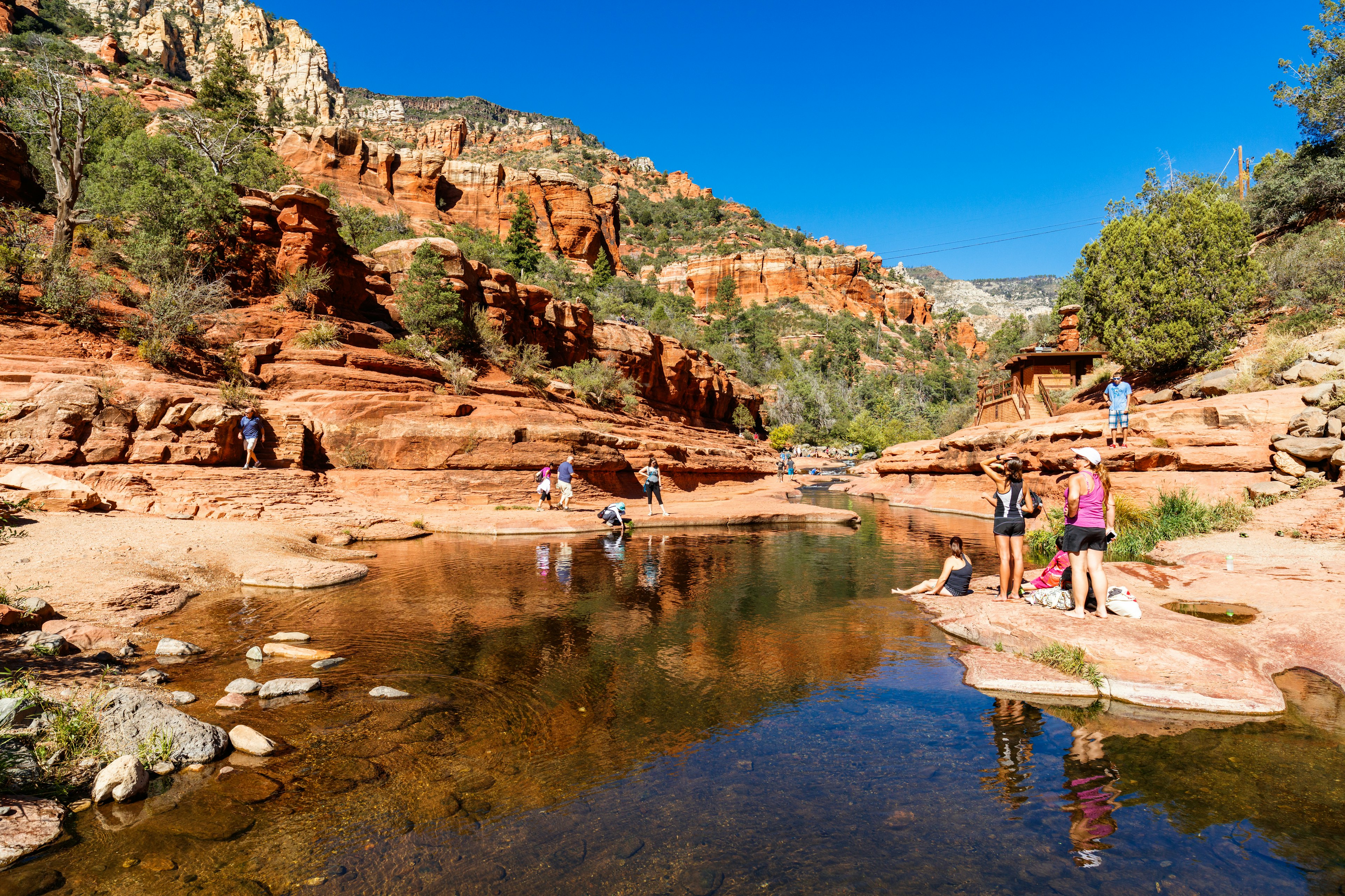 Several groups of people in outdoor clothing linger around a creek at the bottom of a canyon of tall red rocks