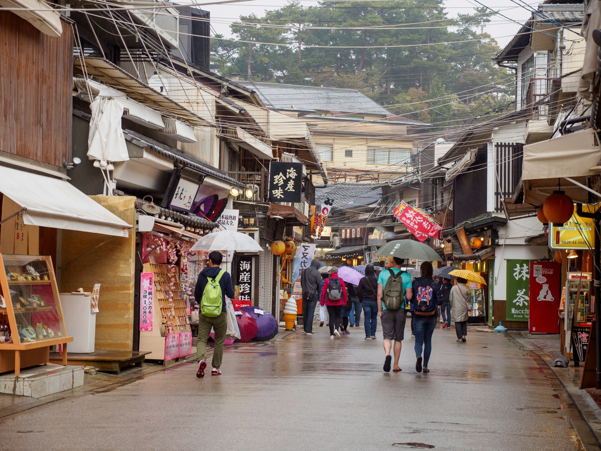 People walk under umbrellas on a street lined with shops on a rainy day