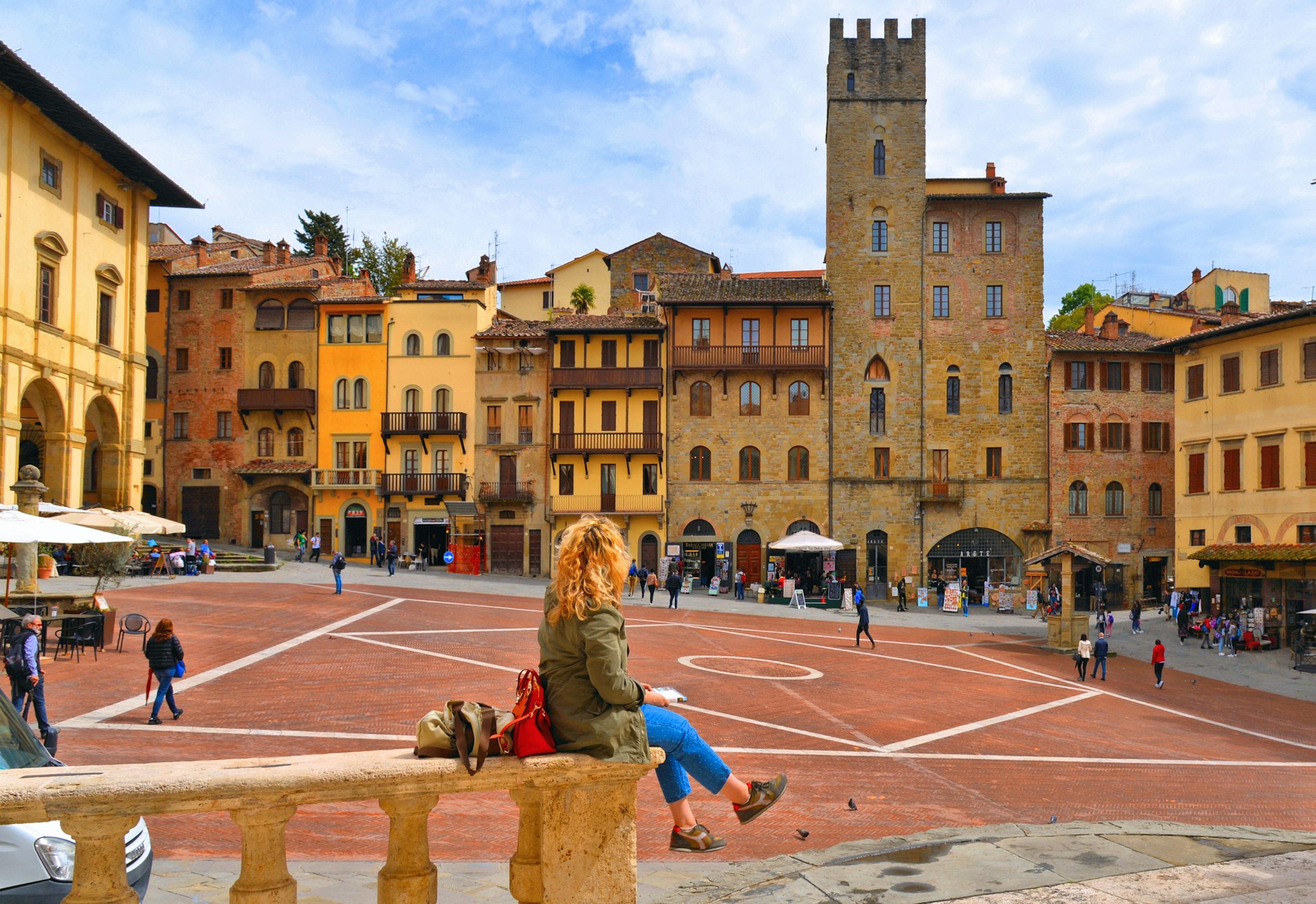 Woman sitting on a stone handrail facing towards a city square dominated by a tower