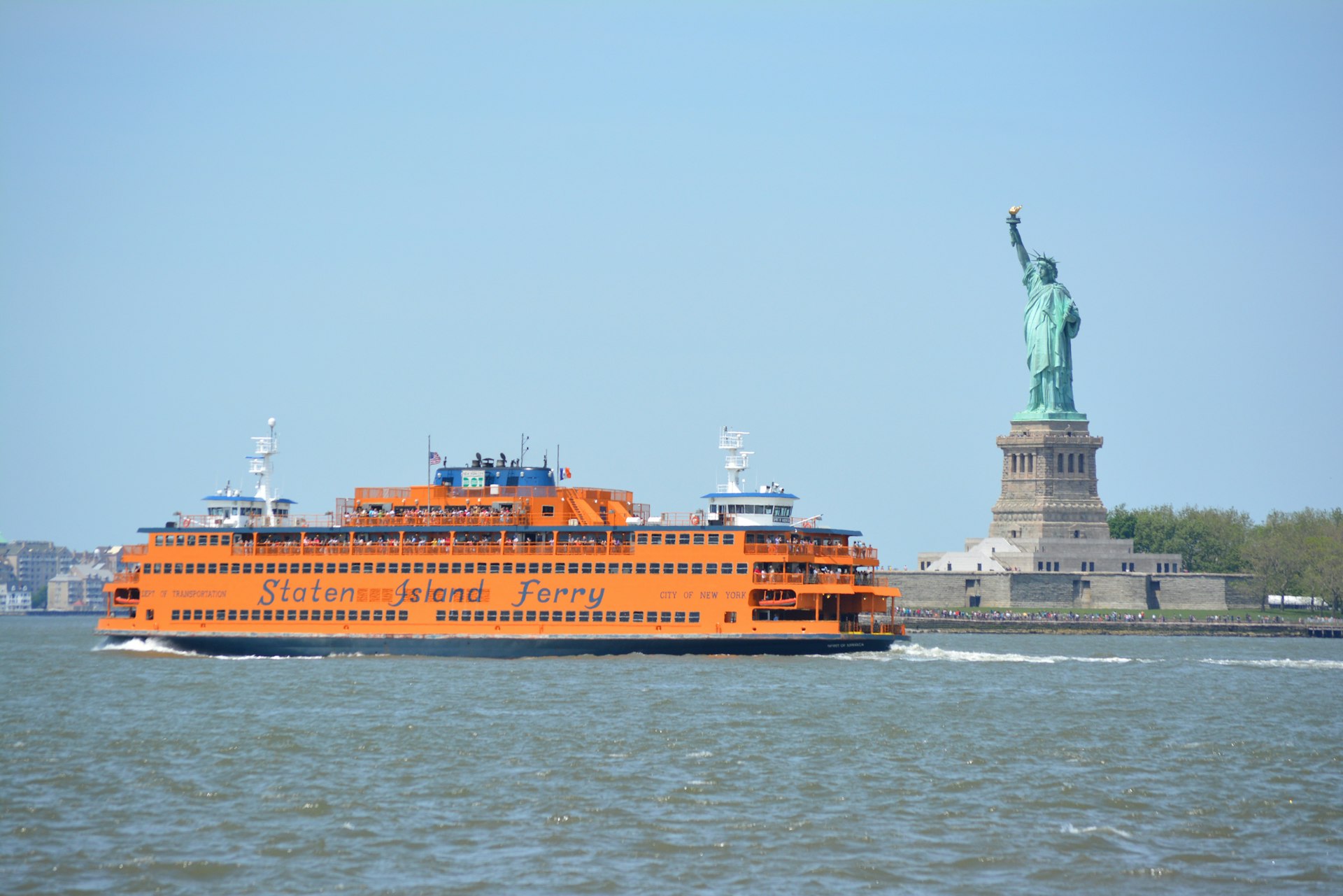 A large yellow-orange ferry sails through a bay with a tall statue of a female holding a burning torch aloft